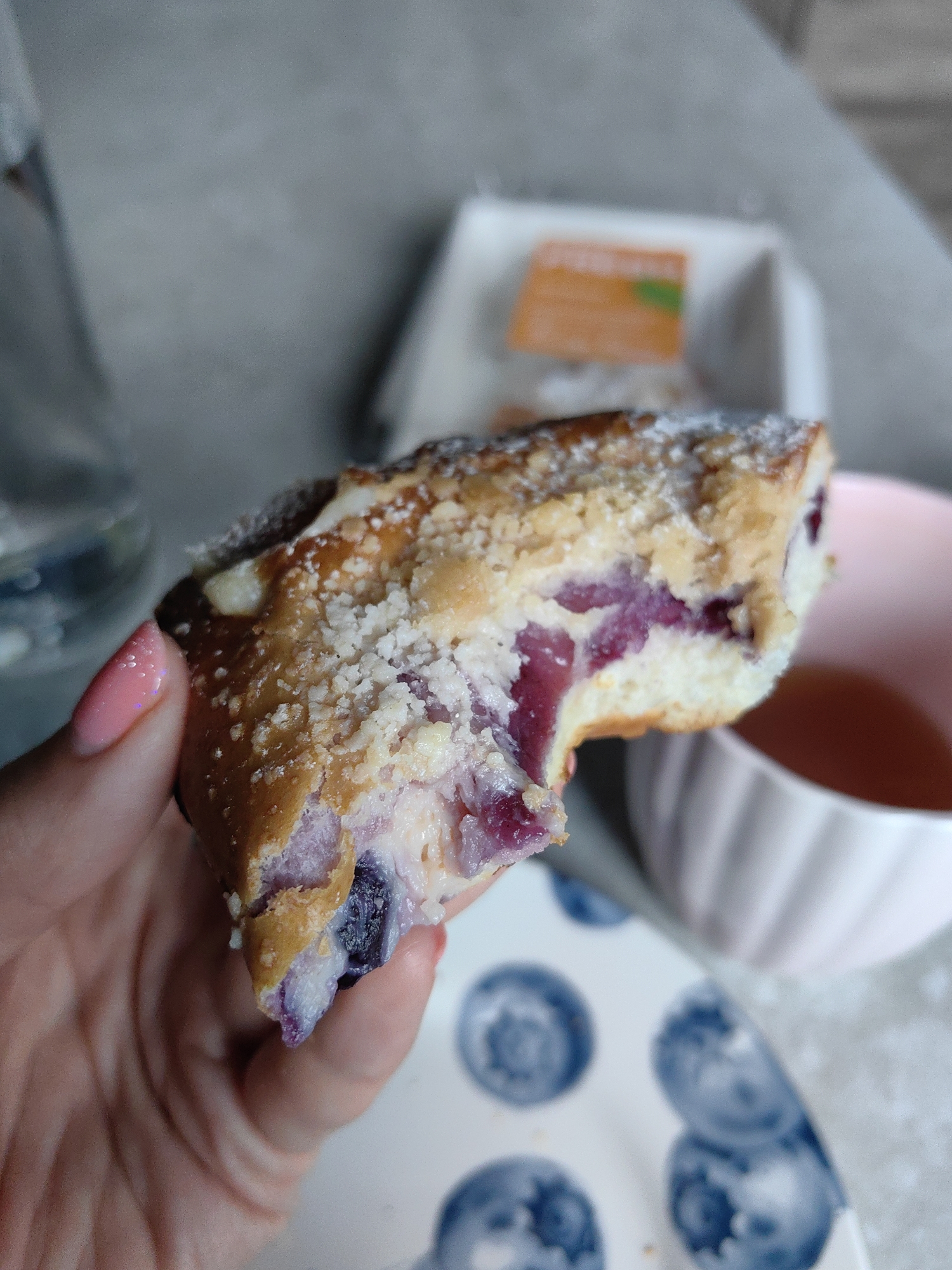 A close-up shot captures a slice of blueberry cheesecake pastry being held by a person's hand. The pastry has a crumb topping and visible blueberries within the cheesecake filling. In the background, a pink teacup with tea, a white dish with blueberries, and a blurry water carafe are visible. The scene appears to be indoors, with a neutral-colored countertop and wall. The image does not provide any information about the location in the city of Fanipal, Belarus.