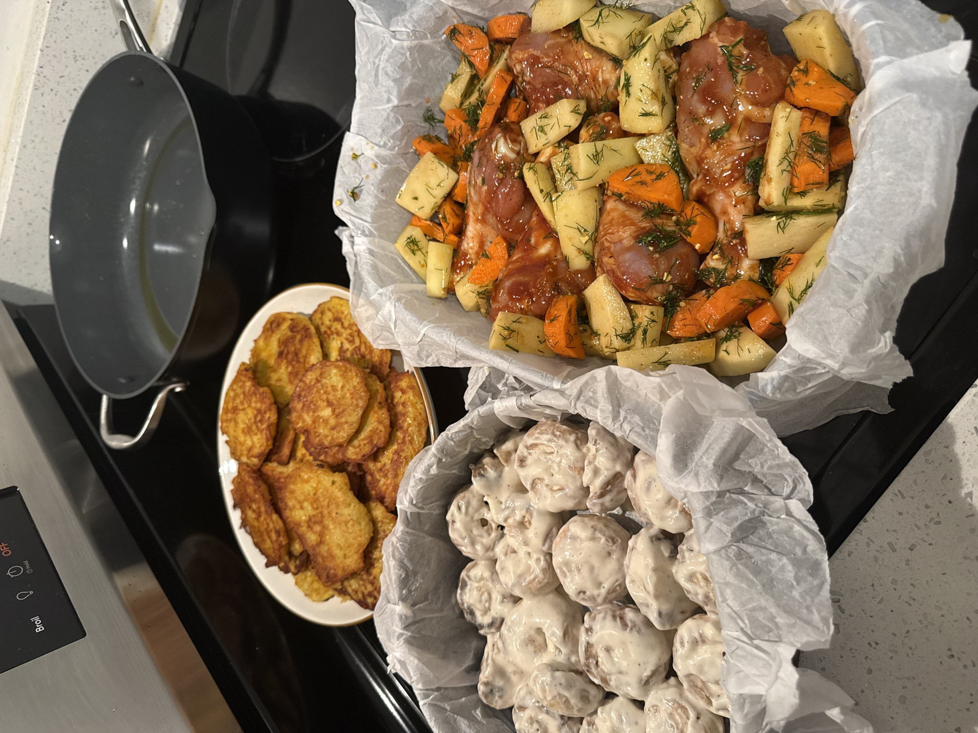 The image shows a kitchen scene with food preparations. In the foreground, two baking dishes are visible. The dish on the left contains numerous small, round, doughy items coated in a creamy sauce. The dish on the right holds pieces of raw meat, possibly chicken, seasoned and interspersed with chopped carrots, potatoes, and dill. Behind these dishes, a plate piled high with golden-brown potato pancakes is placed on a stovetop. To the right of the plate of pancakes, a dark-colored frying pan with a small amount of oil is also on the stovetop. The background shows a stainless steel oven control panel with buttons labeled "Broil," a lightbulb icon, a clock icon, and "Hold," as well as an "OFF" button. The overall environment suggests a home kitchen where a meal is being prepared. The image does not contain any information that allows for the determination of its location in New York City, United States of America.