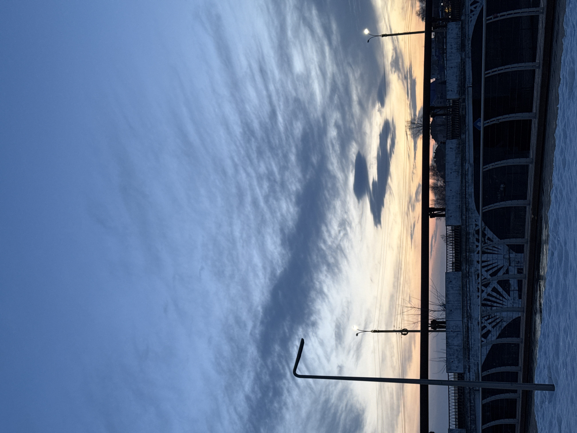 The image depicts an evening sky over the embankment of the Votkinsk Pond in Votkinsk, Russia. Wispy, layered clouds dominate the sky, transitioning from a deep blue in the upper left to lighter hues closer to the horizon. Two lampposts are visible along the embankment. The balustrade of the embankment is on the right, showing its arch details and dark color. Snow is present on the ground.