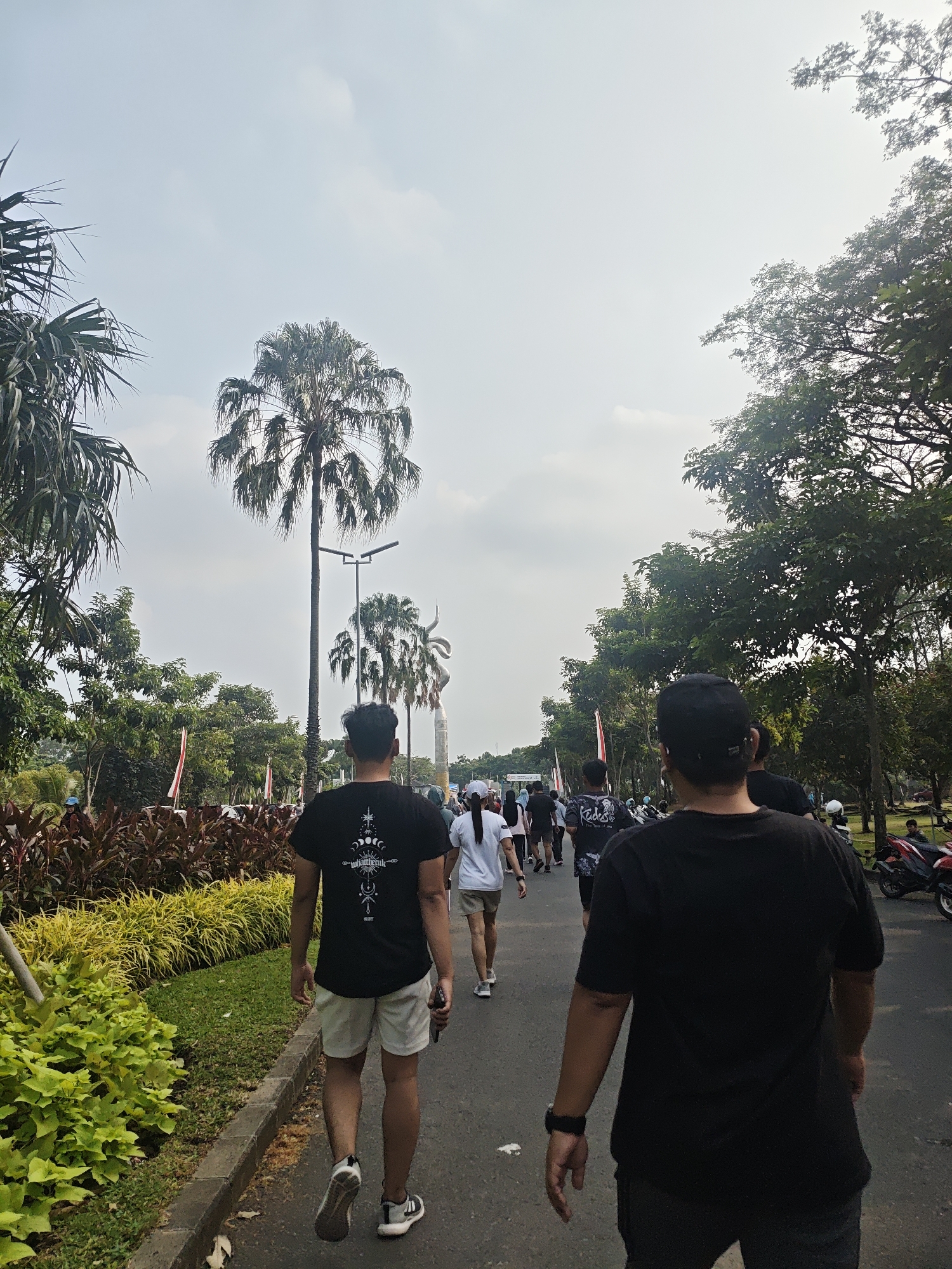 A group of people are walking on a paved road lined with trees and vegetation. Two men in black t-shirts are in the foreground, walking away from the camera. The man on the left is wearing light-colored shorts, while the man on the right is wearing dark pants. There are several other individuals walking ahead of them on the road, some of whom are wearing white t-shirts and shorts. In the background, there are tall palm trees and a statue. Red and white flags are visible along the side of the road. The sky is overcast. The scene appears to be in a park or recreational area, possibly in the city of Bacin, Indonesia.