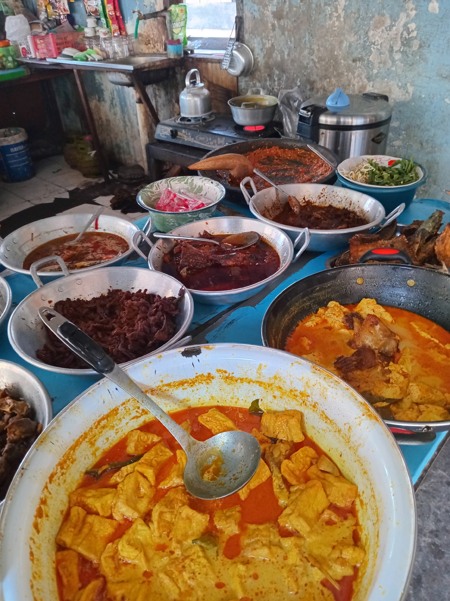 An overhead, eye-level shot captures a vibrant display of Indonesian food, possibly a street food stall in Ngipik, Indonesia. Several metal serving dishes, each containing a different dish, are arranged on a bright blue surface, likely a table. In the foreground, a large white bowl overflows with a rich, orange curry, featuring large chunks of tofu or some other cubed ingredient and a ladle resting within. To the right, a black frying pan holds more of the same curry, with visible pieces of meat or chicken. Behind these, a scattering of fried fish adds textural contrast. Other bowls contain various stews and shredded meats in shades of brown and red, one with a spoon submerged in its saucy contents. A green bowl with white and green speckles holds a small portion of bean sprouts and green beans, suggesting a fresh component to the meal. In the background, a well-worn kitchen area is visible with a propane stove, a metal kettle, and various containers and shelves filled with cooking supplies and food items. The overall impression is one of abundance and a home-style culinary offering.