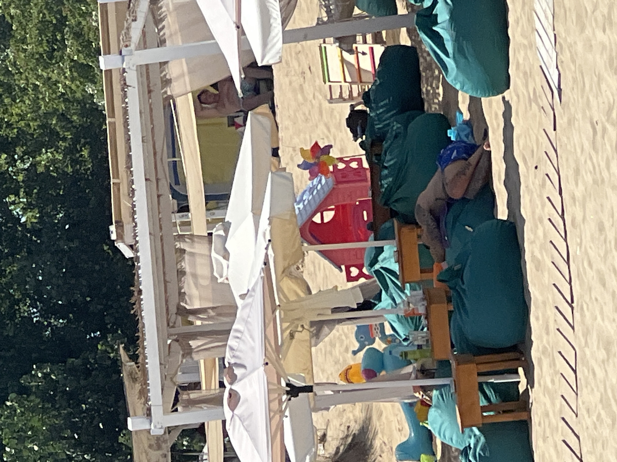 In Sozopol, Bulgaria, a beach scene unfolds under canopies with white fabric roofs. In the background, a person reclines on a chair, partially visible behind a curtain. In the foreground, several large, teal beanbag chairs are scattered on the sand, some occupied by a person lying down with their face down, wearing blue swimwear and having tattoos. A small wooden table is positioned in front of one beanbag. Scattered toys, including an inflatable airplane and an elephant, are visible on the sand, along with a pink playhouse with a colorful pinwheel. The sand is a light beige, and there are diagonal shadows cast on it, suggesting direct sunlight. Trees form a dark green backdrop above the canopies.