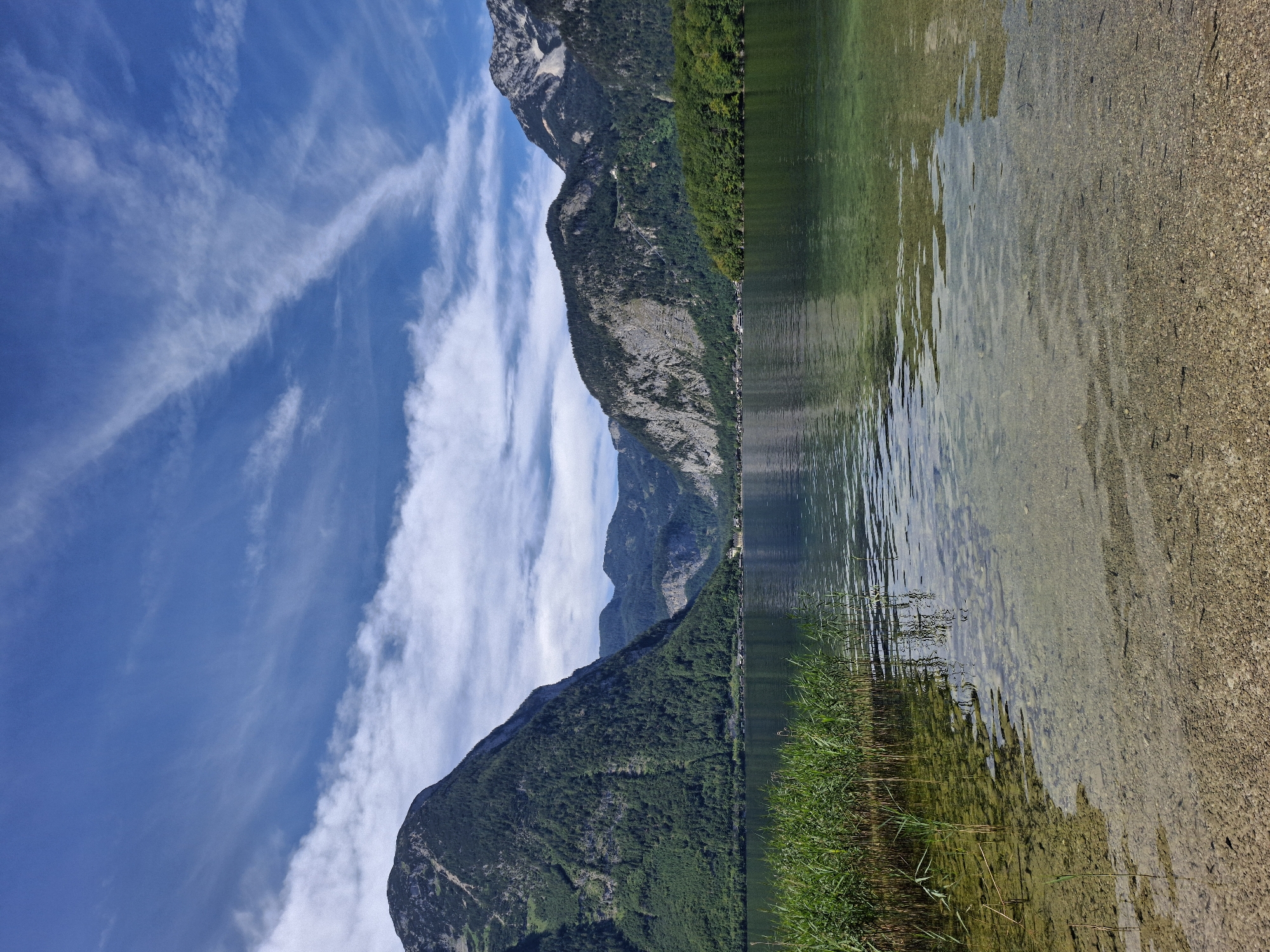 A serene landscape photo depicts a clear, calm lake with a pebbled bottom visible in the foreground. Tall reeds line the left bank, their reflections shimmering on the water's surface. In the background, a dense green forest blankets the slopes of majestic mountains, which rise steeply on either side of the lake. A small village is nestled along the far shore, with houses dotting the landscape. The sky above is a vibrant blue, streaked with wispy white clouds. This scene is characteristic of the natural beauty found in Obertraun, Austria, particularly along the shores of Lake Hallstatt.