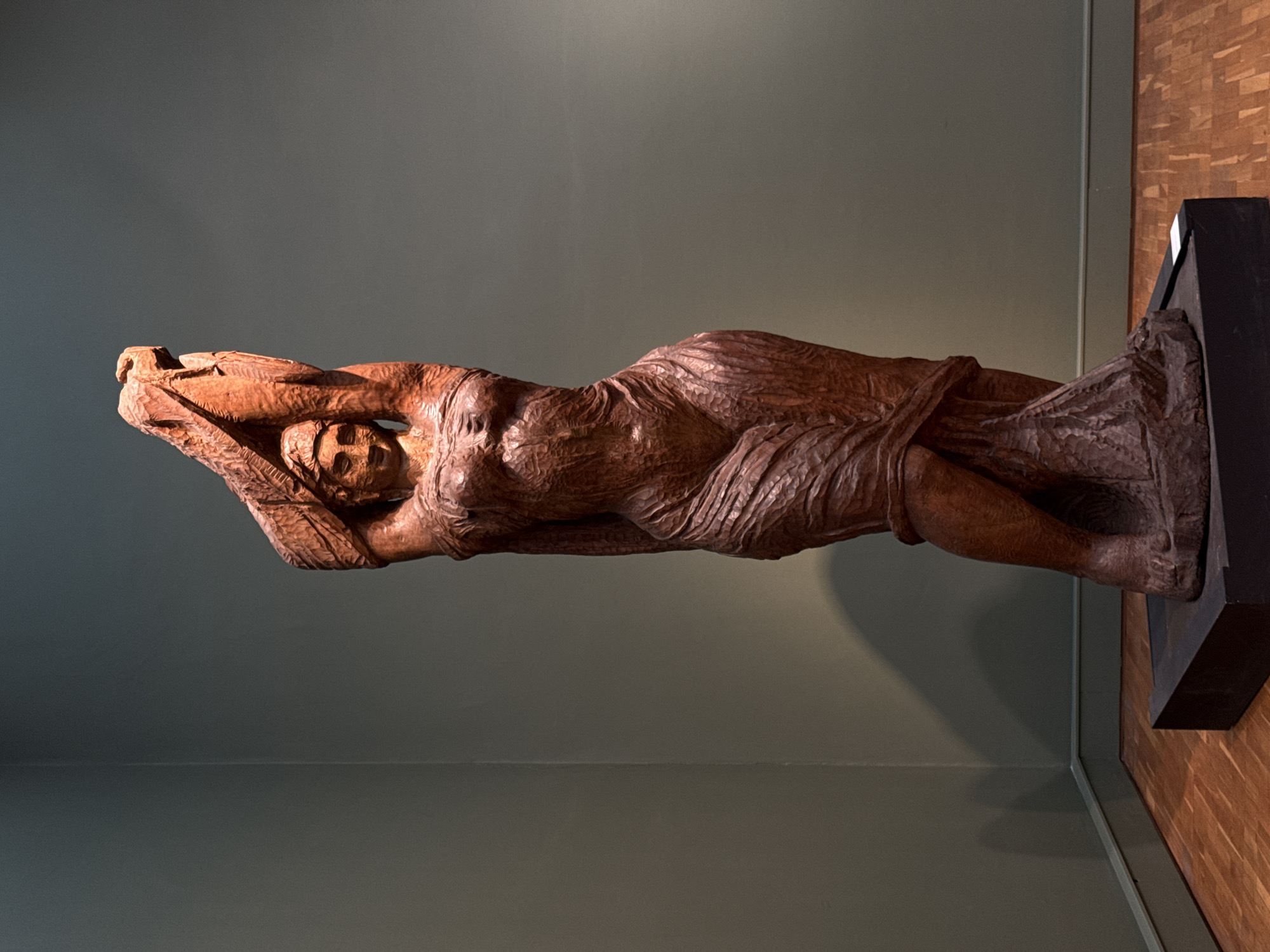The image shows a wooden sculpture of a reclining figure. The figure appears to be a woman, draped in a textured cloth or garment. The sculpture is mounted on a dark base. The background consists of a plain, green-gray wall. To the right, a portion of a wooden wall is visible. The sculpture's location is unspecified within Moscow, Russia, based solely on the provided image.