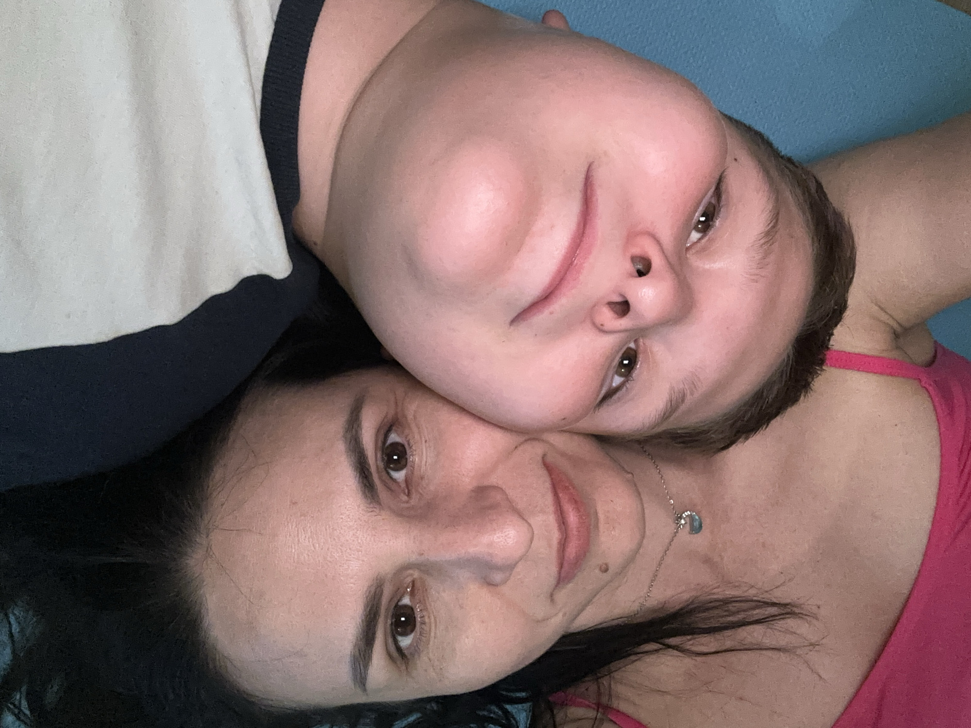 Two people are lying down close to each other. The person on the bottom has dark hair and is wearing a pink tank top and a necklace. The person on top has short brown hair and is wearing a white and dark blue top. The location is Unknown, Unknown.