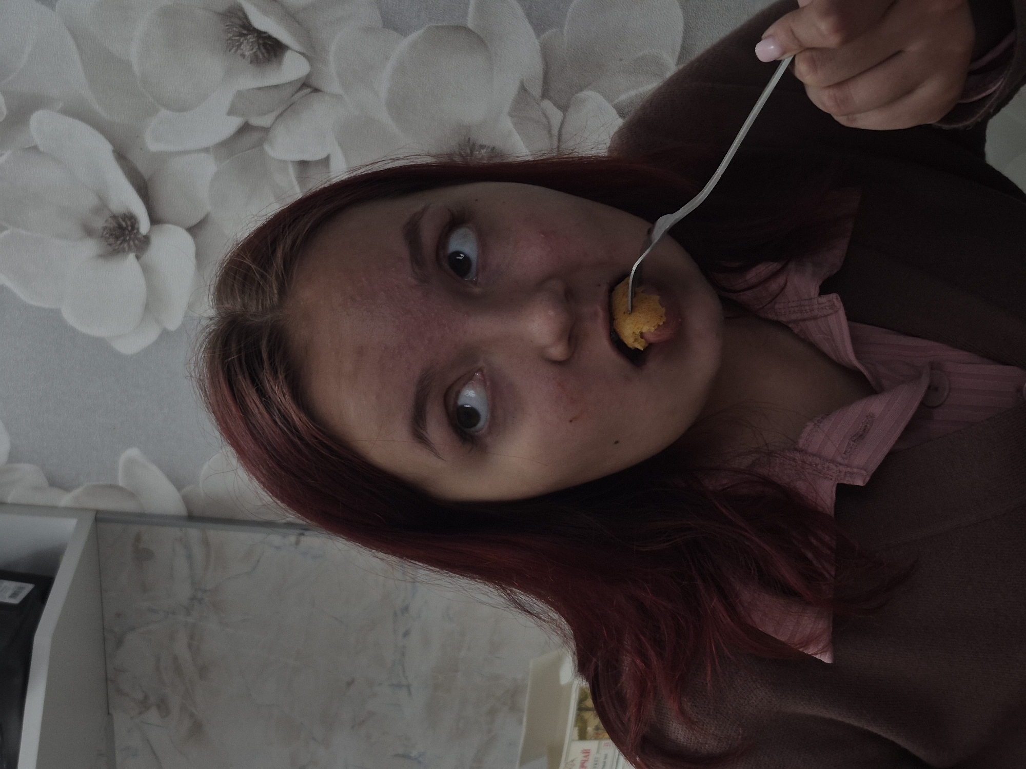 A young woman with reddish-brown hair is holding a fork with a piece of food up to her mouth. The woman is wearing a pink button-down shirt and a brown sweater. The background includes a wall with a floral pattern and a partial view of a kitchen cabinet. The photograph may have been taken in Chaikovsky, Russia, but this cannot be determined from the visual content alone.