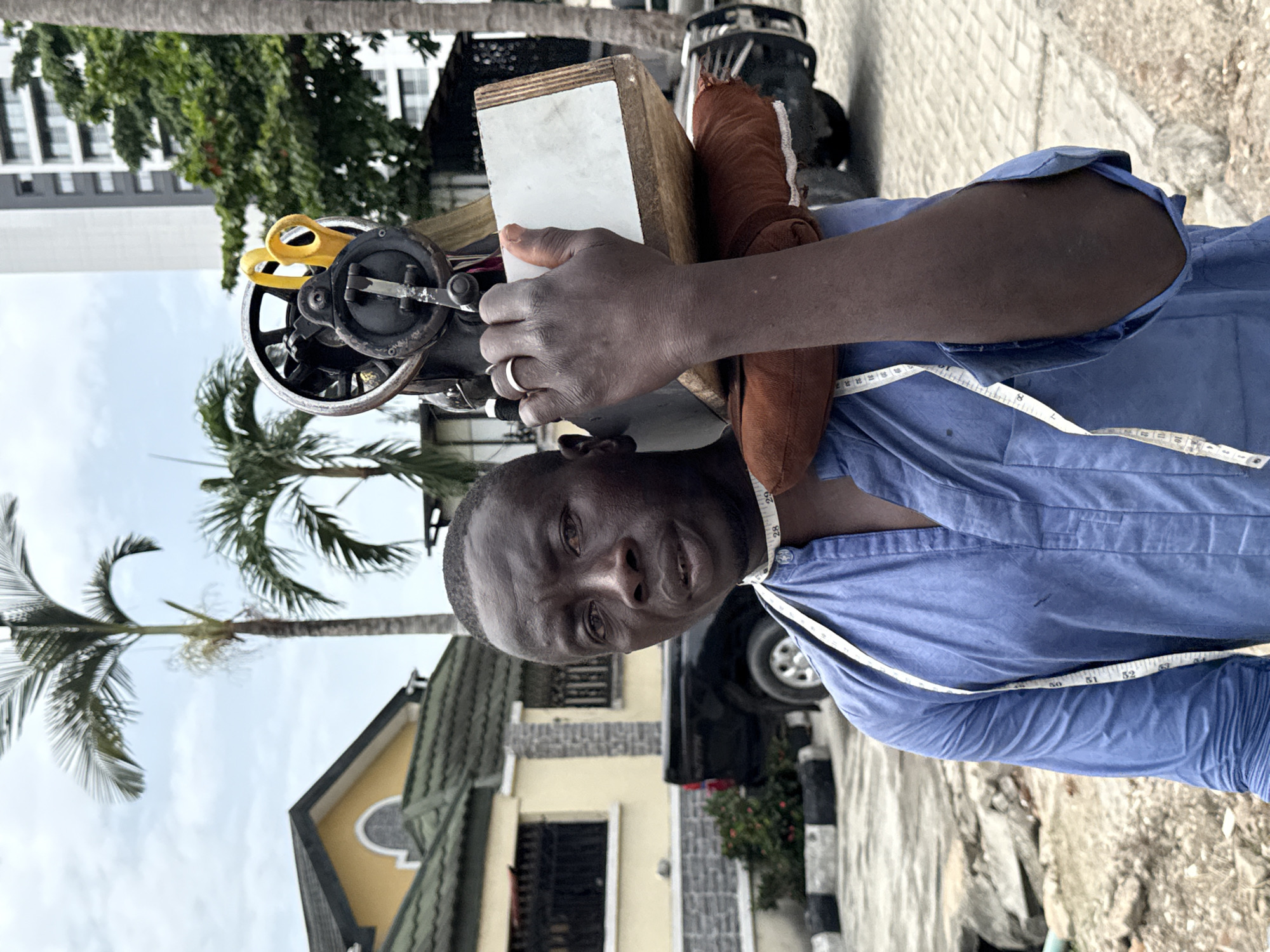 In Itirin, Nigeria, an adult man is captured on a city street carrying a sewing machine on his shoulder. The man, with dark skin and short hair, wears a blue collared shirt and a white measuring tape around his neck. He is also wearing a ring on his left hand. On his shoulder is a sewing machine and its wooden box. A pair of yellow scissors are visible near the sewing machine. In the background, there is a yellow and grey house with a dark door, a black vehicle, trees, and a multi-story building. The ground is paved.