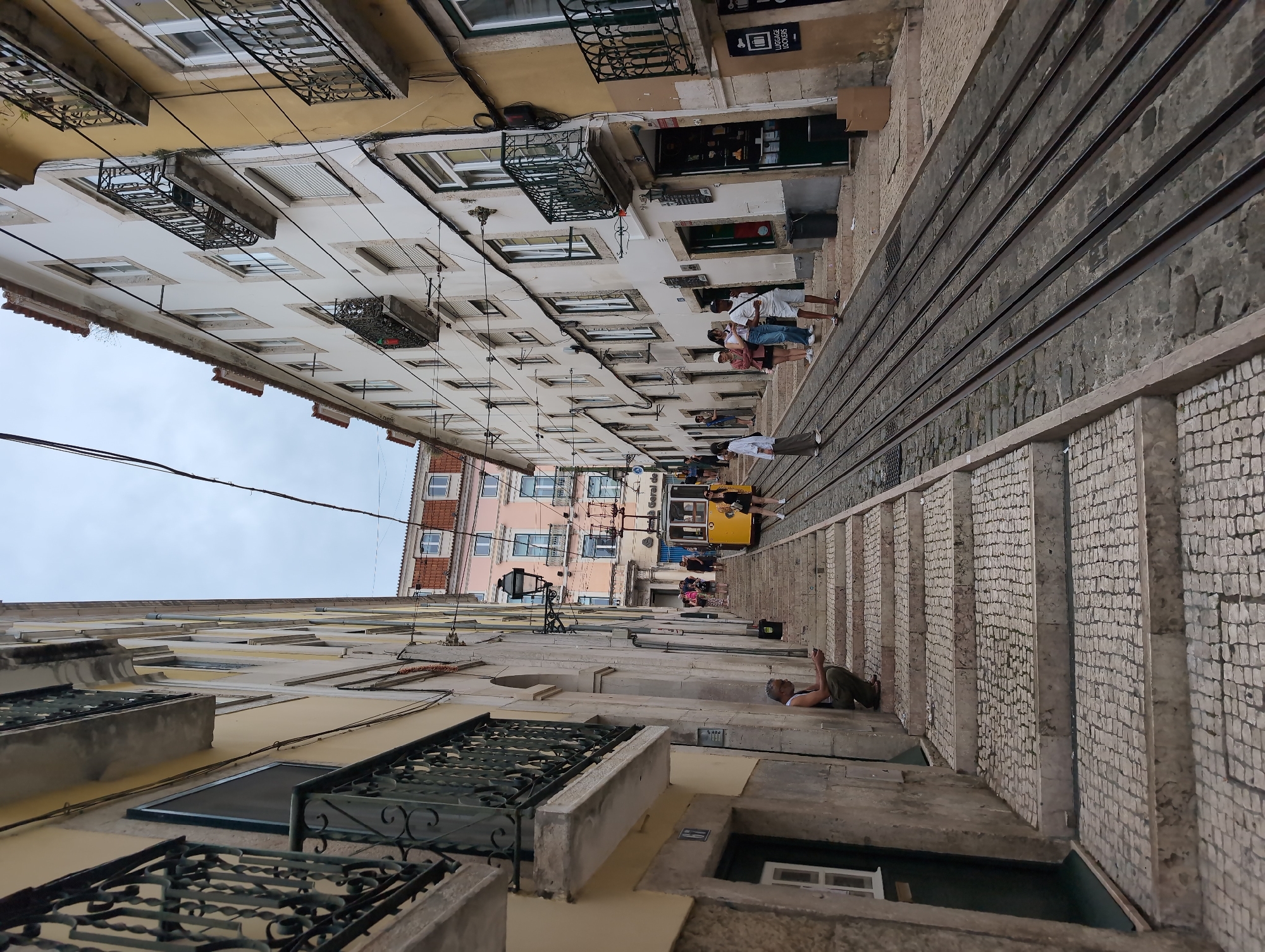 The image shows a street view in Lisbon, Portugal, featuring the steep incline and tram tracks of the Ascensor da Bica funicular. Buildings line both sides of the street, with varied architectural styles and colors. Balconies with intricate ironwork are visible on some buildings. People are present, some standing on the steps alongside the tram tracks and a few in the background, near a yellow tram. One man is resting on the steps. The cobblestone paving adds to the historic atmosphere of the location. Overhead power lines are visible.
