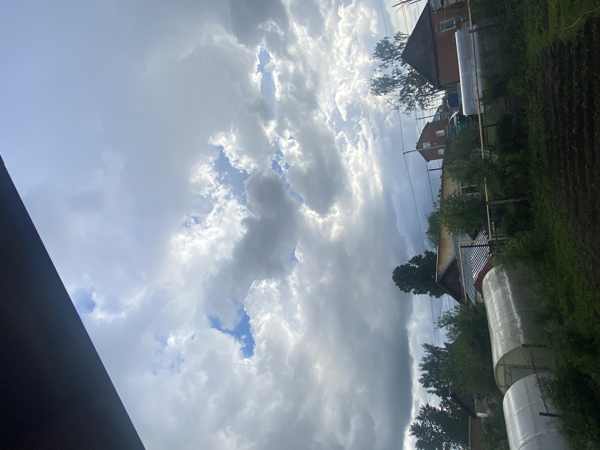 This media file captures a rural or semi-rural landscape under a dynamic sky. The scene is dominated by a partially cloudy sky, featuring large, puffy cumulus clouds in various shades of white and grey, interspersed with patches of clear blue sky where sunlight breaks through.
In the middle and background, several buildings are visible, typical of a village or dacha setting. There are residential-style houses with sloped roofs, one with a dark red roof and brick walls, and others partly obscured by trees. The houses are surrounded by lush green foliage, including deciduous trees and bushes.
Prominently featured in the midground are at least two large, tunnel-shaped greenhouses with translucent coverings, commonly used for gardening. Adjacent to these, sections of cultivated land with dark soil and sparse green growth are visible, suggesting garden beds. Utility poles and power lines can be faintly seen against the sky.
A dark, angular structure, possibly a roofline or part of a building, is present in the extreme foreground on the left, partially framing the upper left portion of the sky.
The time of day appears to be daytime, likely late morning or afternoon, given the bright, yet partly overcast, conditions. The weather is characterized by scattered clouds. The presence of greenhouses and garden plots aligns with the provided location context of Chaikovsky, Russia, suggesting a typical countryside environment. No people or visible text are present in the image.