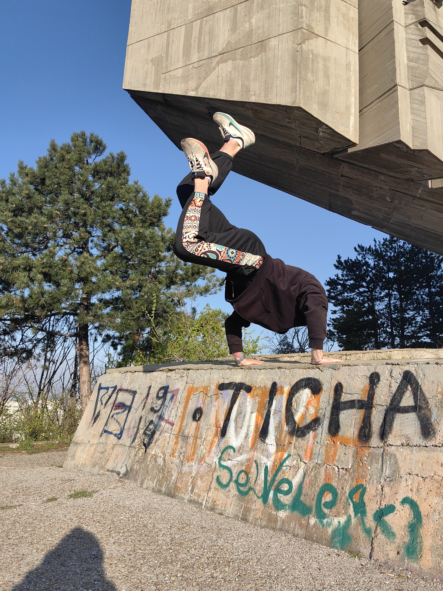 A person in a dark hoodie and patterned sweatpants is performing a handstand on a concrete wall with graffiti. The graffiti includes the word "TICHA" in black paint and "SeVeLeasT" in green. The person is wearing sneakers. Above them is a large concrete structure. In the background, there are trees and a clear blue sky. The location is in Varna, Bulgaria, near the Monument of the Bulgarian-Soviet Friendship.