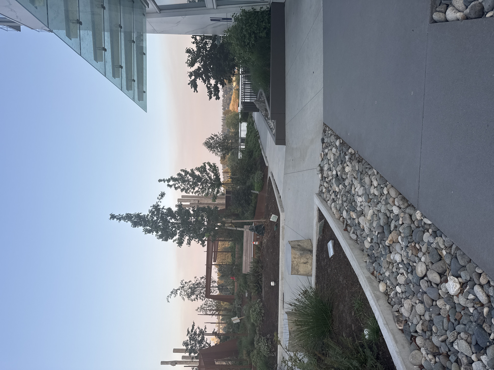 This is a medium-shot, eye-level, vertical photograph of an outdoor common area adjacent to a modern building. In the foreground, a dark gray paved walkway runs diagonally across the frame from the bottom right corner. To the left of the walkway, a raised garden bed is bordered by a concrete edge and filled with dark soil, lush green plants, and a decorative arrangement of smooth, gray and white river stones. Further back, two large, rough-hewn stone stumps serve as seating. A pathway of light gray concrete extends into the middle ground, curving to the left. Beyond this pathway, a well-maintained garden features various plants, shrubs, and trees, along with wooden structures, a bench, and what appears to be a small pond or water feature. In the background, a body of water, possibly a river or lake, is visible, with a distant treeline and hills under a pale, hazy sky. The building on the right side of the image features a white facade with large windows and a distinctive, angled glass canopy extending from the roofline. The scene is devoid of people and appears to be a public or shared space, possibly in a residential or commercial development. The lighting suggests it is either early morning or late afternoon, with soft, diffused light. The presence of hazy sky may indicate smoke from wildfires. This scene is located at the River District development in New Westminster, Canada.