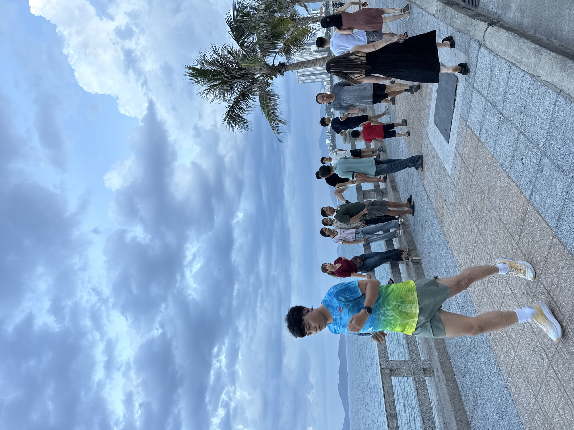 The image depicts a coastal promenade in Nha Trang, Vietnam, under a cloudy sky. A young man is captured mid-stride, jogging along the paved walkway. He wears a blue and green tie-dye t-shirt and olive green shorts, white socks, and athletic shoes.
To his left, the ocean stretches out, bordered by a concrete railing. A line of people are visible further down the promenade, some standing and conversing, while others walk. Among them are several women and men of various ages, dressed in casual attire. Behind the people, palm trees and modern high-rise buildings form the urban backdrop. The promenade itself is paved with square tiles in shades of beige and grey, with a darker rectangular grate set into the ground. The overall atmosphere appears to be that of a public space bustling with activity during a late afternoon or early evening.