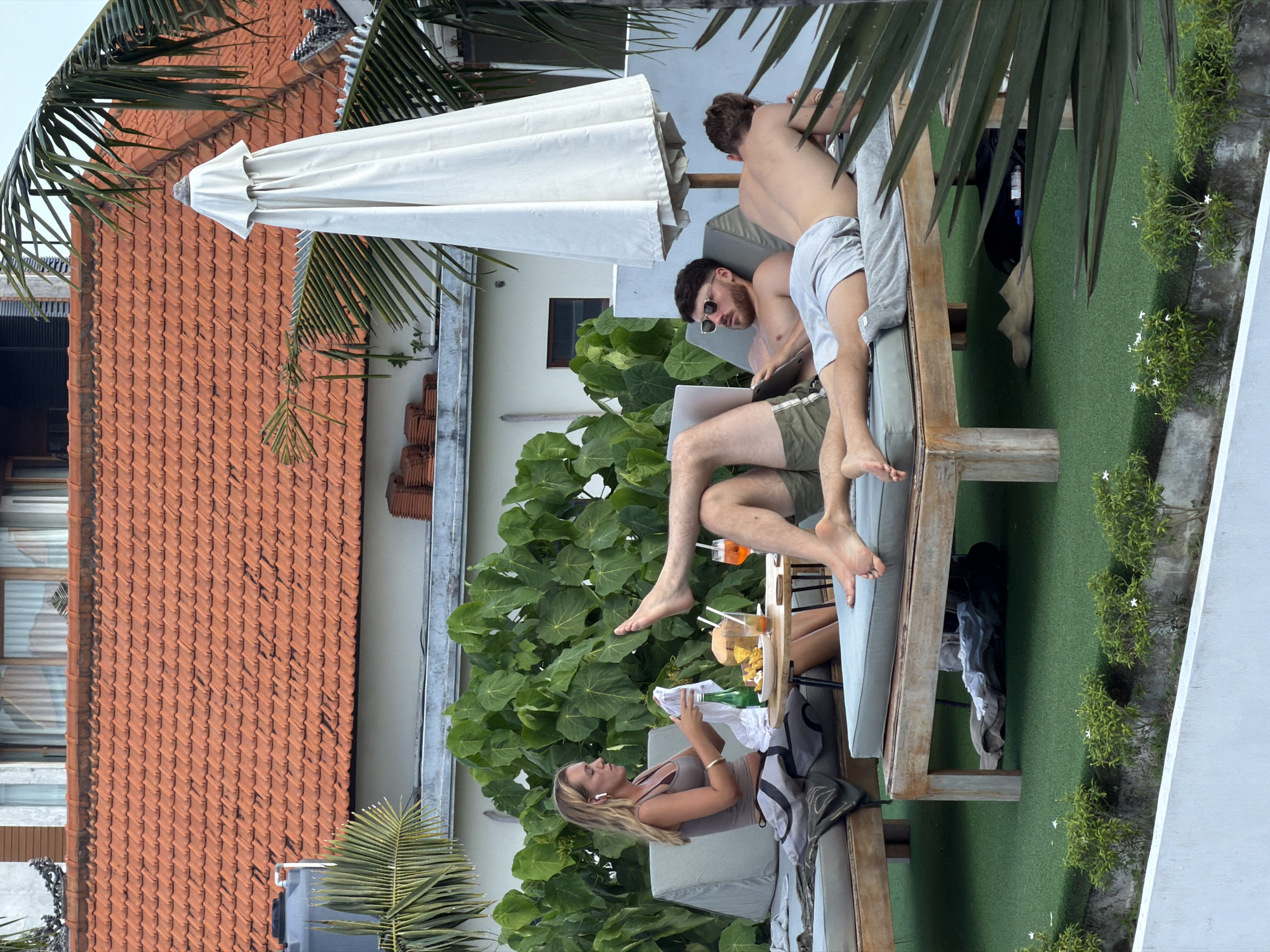 Three people are relaxing on lounge chairs on a rooftop terrace. A woman with blonde hair is sitting up, holding a white cloth and a glass, with a drink and snacks on a table in front of her. Two men are lying on lounge chairs. One man is lying on his back, wearing sunglasses and looking at a laptop. The other man is lying on his stomach, facing away from the camera. The terrace has green turf flooring and is surrounded by lush green foliage. In the background, a building with a red tiled roof is visible. The scene appears to be in a resort or hotel in North Kuta, Indonesia, likely a poolside or sunbathing area given the lounge chairs, drinks, and tropical vegetation.