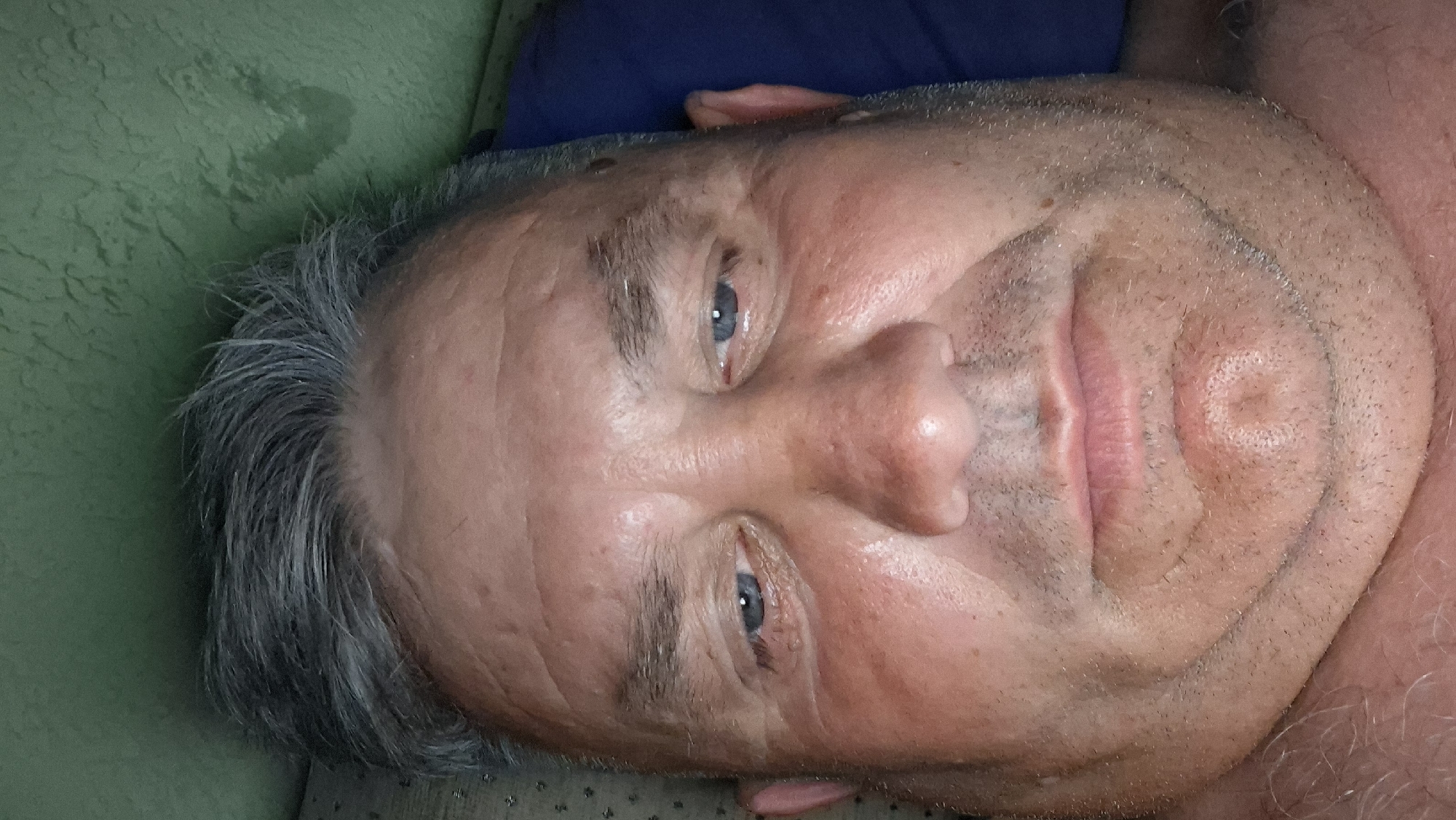 A close-up, front-facing selfie of a middle-aged Caucasian man with graying hair and blue eyes is shown. He is lying down, possibly in bed, with a green wall and a dark blue pillow visible in the background. The lighting suggests it is nighttime or a dimly lit room. The man appears to have a full face and a double chin, with some stubble on his chin and upper lip. His skin has a slightly red, flushed tone, and there are visible pores and some wrinkles on his forehead and around his eyes. He is looking directly at the camera with a neutral expression. No specific location in Voinova, Moldova, can be identified from the image.