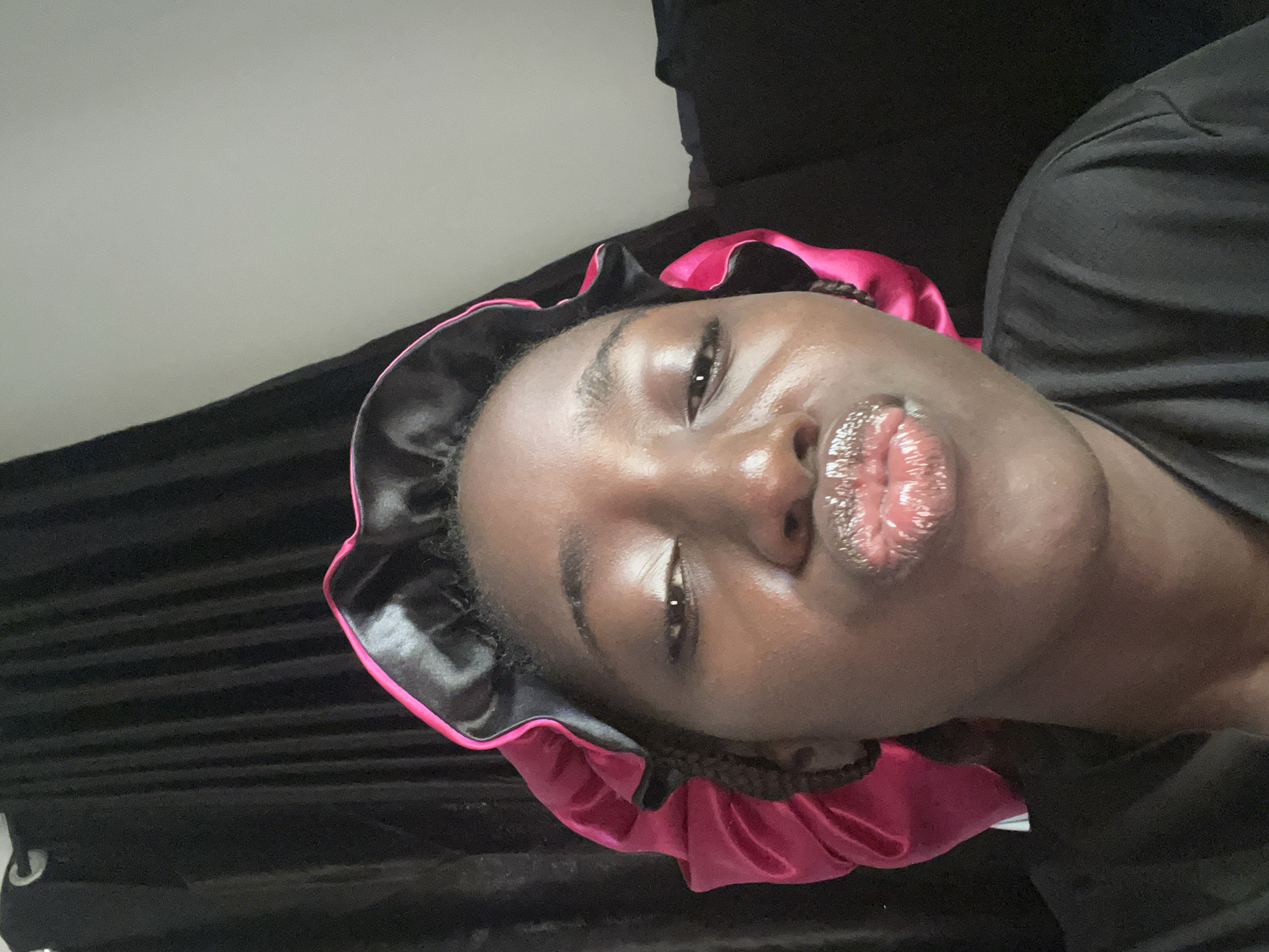 A close-up, rotated selfie of a Black woman wearing a black and pink satin bonnet and a black shirt. The background consists of a light-colored wall on the left and a dark, draped fabric behind her. The location is not discernible as Abuja, Nigeria.