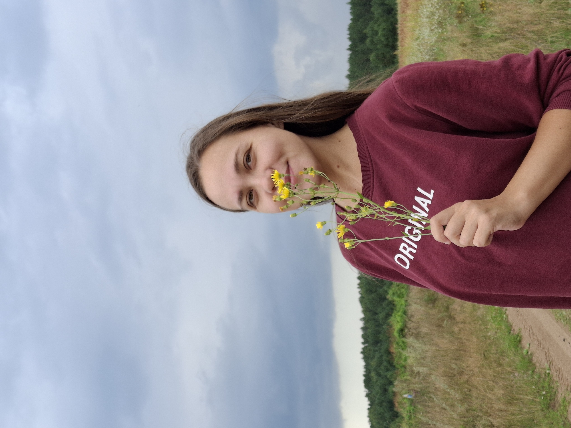 A young woman with long, brown hair stands in a field in Plishkino, Russia, holding a small bouquet of yellow wildflowers up to her face. She is wearing a maroon sweatshirt with the word "ORIGINAL" printed on it in white. The background features a dirt path, dry grass, a line of green trees, and a cloudy sky.