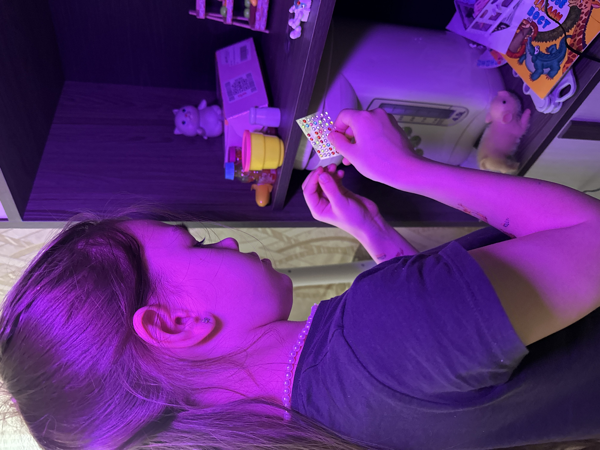 The image captures a person, likely a young female, in an indoor setting illuminated by strong purple/magenta artificial light. She is viewed from a side profile, looking down and focused on a small sheet of colorful, faceted adhesive gems or stickers held in her hands. Her left hand holds the sheet, while her right hand appears to be either peeling off a sticker or examining them closely.
She has long, light-colored hair, which is also tinted purple by the lighting, and wears a dark t-shirt along with a light-colored, possibly pearl, beaded necklace. A small, sparkly stud earring is visible in her left ear.
The background features a dark wooden shelving unit containing various objects. On the left shelf, a light purple toy figurine resembling a pig or bear is visible, alongside a yellow container with a pink lid, and other small items. On the right shelf, a light brown or beige hamster-like figurine is present, along with a children's book displaying colorful illustrations of animals such as a giraffe and a dinosaur. Partially visible Cyrillic text on the book reads "BОСТ". Another item on the shelf appears to be a white box with a QR code and some indistinct text.
The prominent purple lighting creates a distinct ambiance, suggesting an evening or nighttime setting with mood lighting. The individual appears engrossed in the activity of handling the decorative stickers.