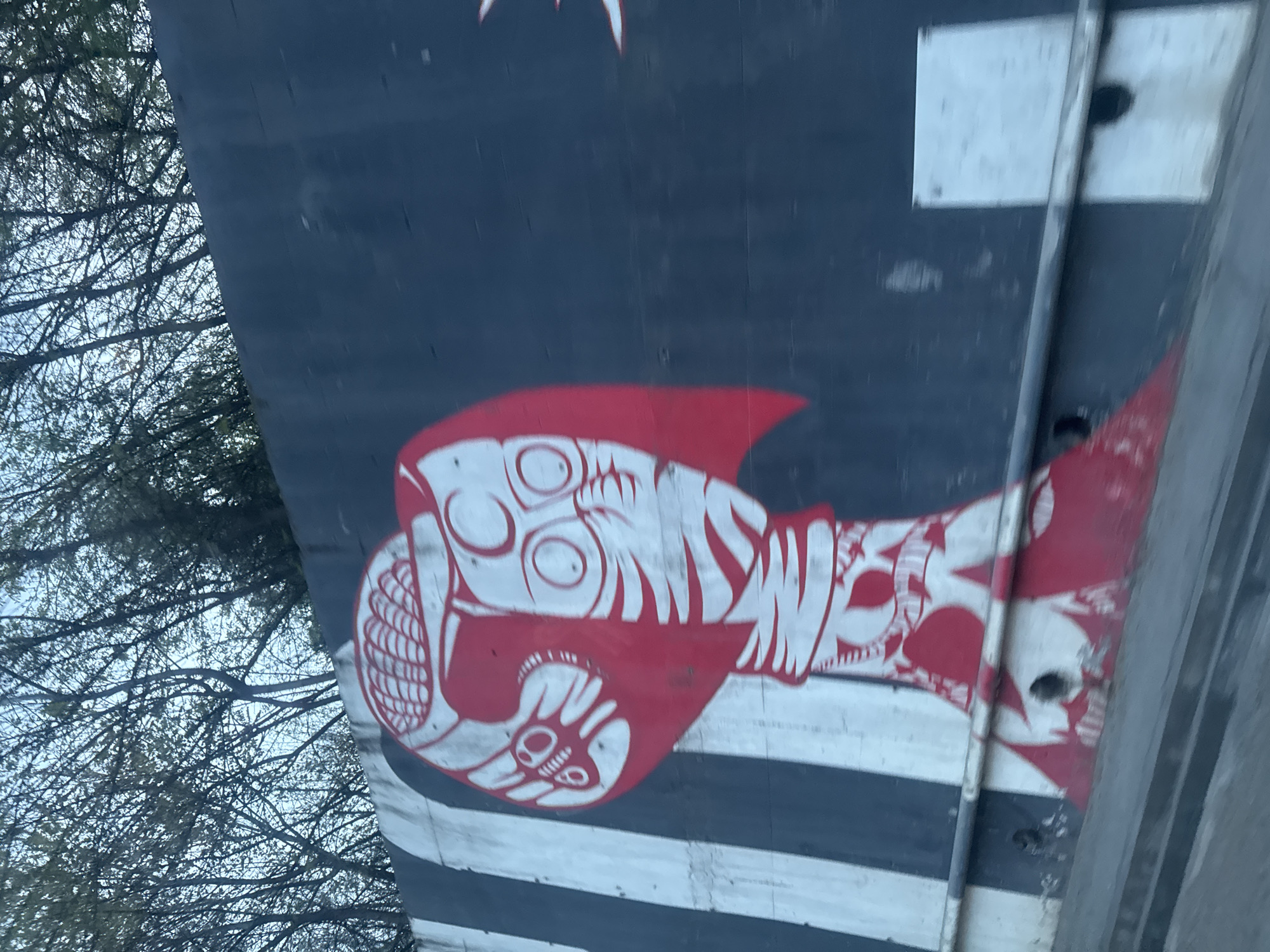 The image shows a portion of what appears to be the tail section of an aircraft, featuring a red and white design. The design resembles a stylized fish or creature with prominent eyes and teeth. The background is a dark blue color with white horizontal stripes. The tail section is mounted on a vehicle or structure. Branches of trees are visible in the upper left corner of the frame, indicating the scene is outdoors. The location is in the city of Tbilisi, Georgia.