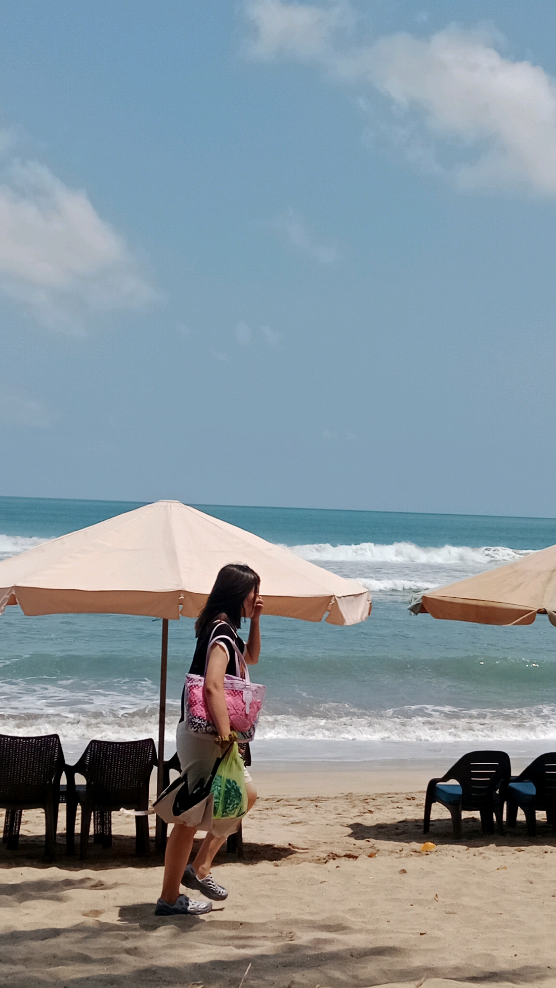 A woman walks along the sandy beach in Kuta, Indonesia, with the turquoise ocean and crashing waves visible behind her. She carries a pink patterned tote bag and a green plastic bag. Two large, light-colored umbrellas provide shade on the beach, and several dark-colored chairs are visible in the foreground and background. The sky is a clear, bright blue with scattered white clouds. The scene depicts a sunny day at a popular tourist destination.
