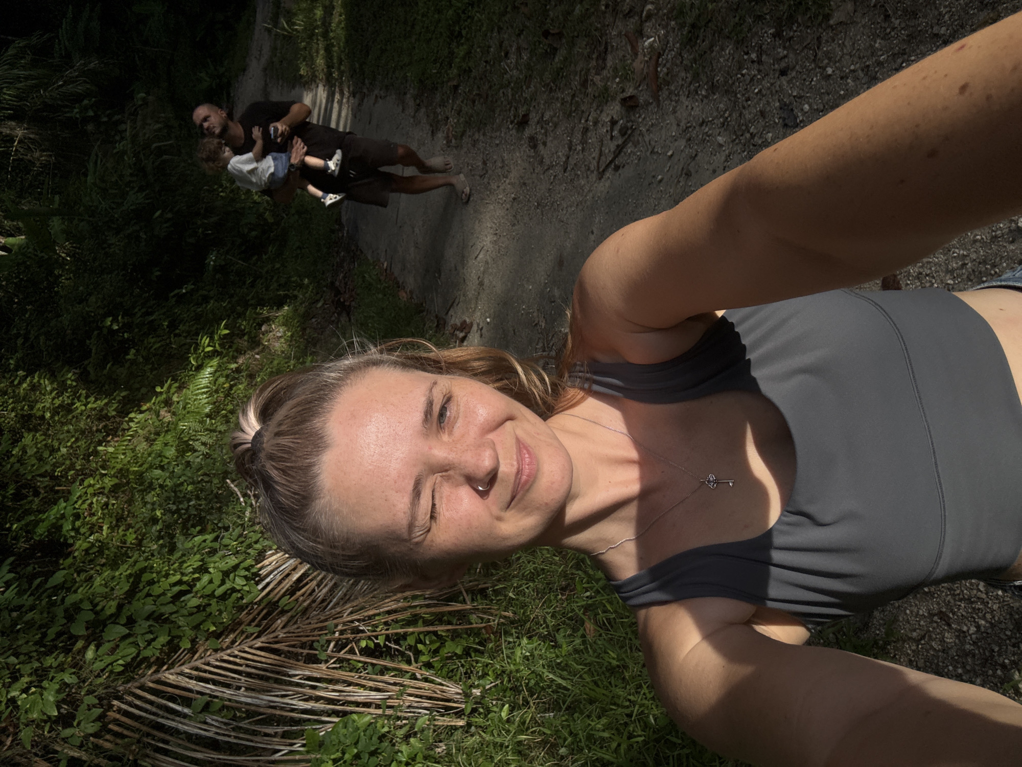 In a sun-dappled jungle path, a woman in a grey sports bra takes a selfie, winking at the camera. Behind her, a man carries a young child, both walking away from the viewer. The path is made of gravel and dirt, flanked by lush green foliage. Sunlight filters through the leaves, casting shadows and highlighting the textures of the jungle. The woman's blonde hair is pulled back in a messy bun, and she wears a silver necklace with a key pendant. The man is dressed in a black t-shirt and shorts, while the child wears a white t-shirt and blue shorts. The overall scene depicts a casual outdoor outing in a natural environment.