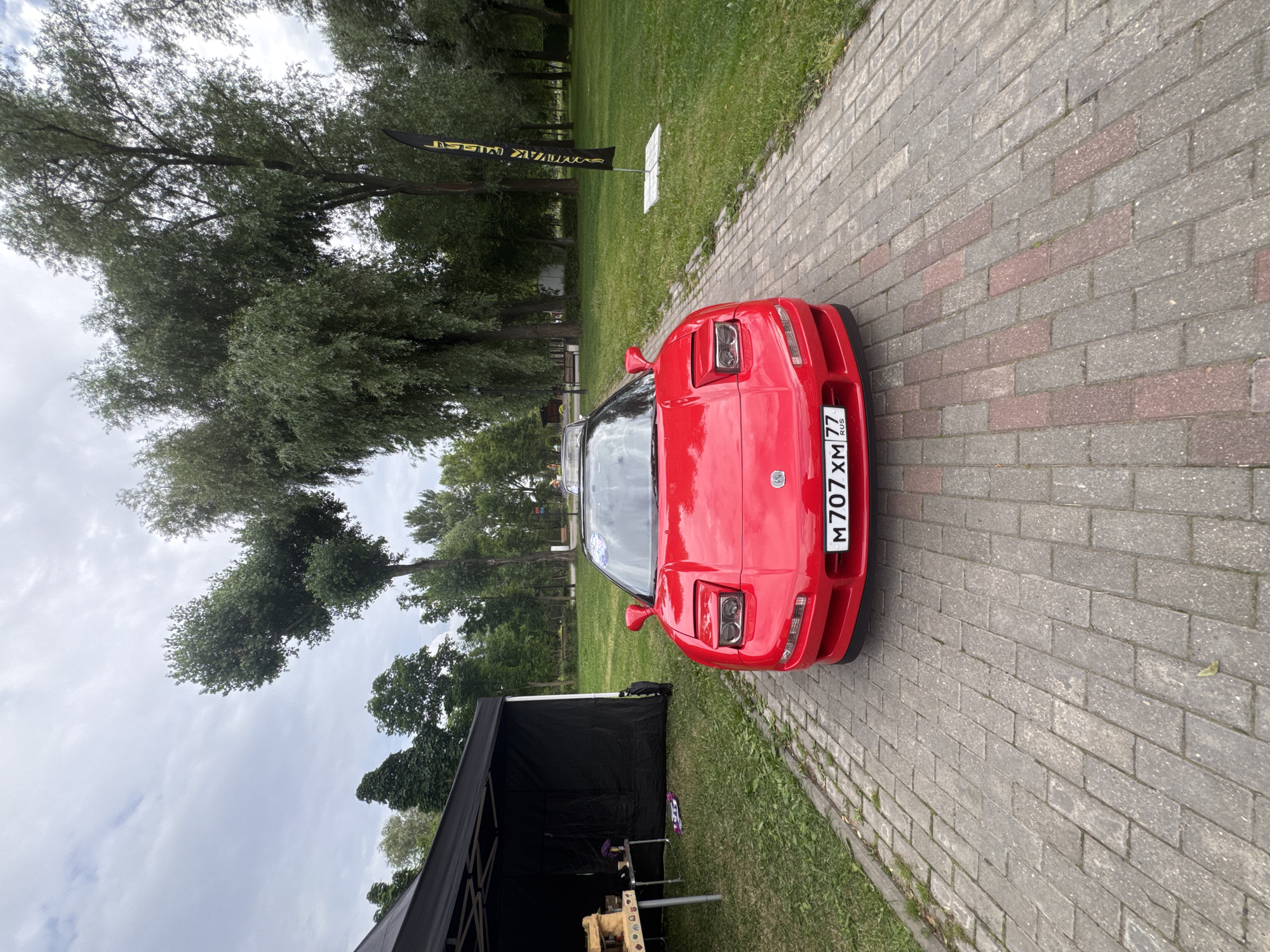 The image displays a red Acura NSX sports car parked on a brick-paved pathway outdoors. The car's headlights are visible, and its license plate reads "M 707 XM 77 RUS". To the left of the car, there is a black tent with a table set up inside, displaying what appears to be a model or display. In the background, there is a grassy area with trees, and a black and yellow banner is planted in the ground. The sky is overcast. The scene appears to be at an outdoor event, possibly a car show or gathering. The presence of Russian license plates and text on the banner suggests the location is within Russia. While the specific location in Moscow is not explicitly stated, the overall ambiance points to an urban park or event space. No people are clearly visible in the image.