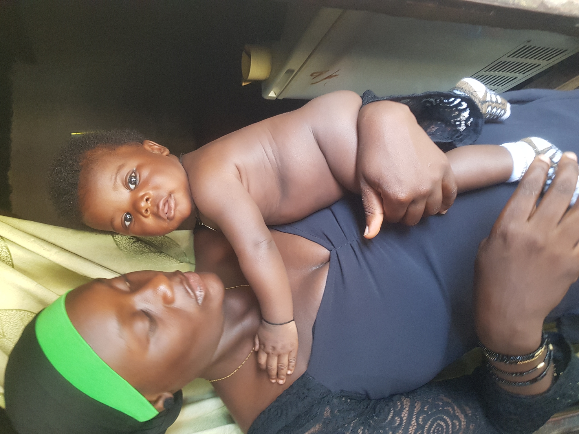 A dark-skinned mother in a black dress with lace sleeves holds her dark-skinned baby. The mother wears a black headscarf with a green band. The baby, wearing white socks with brown and white stripes, is held against the mother's chest. The baby's right arm is draped over the mother's shoulder. The background shows a light-colored curtain and a beige refrigerator with a yellow cup on top. The scene is indoors.