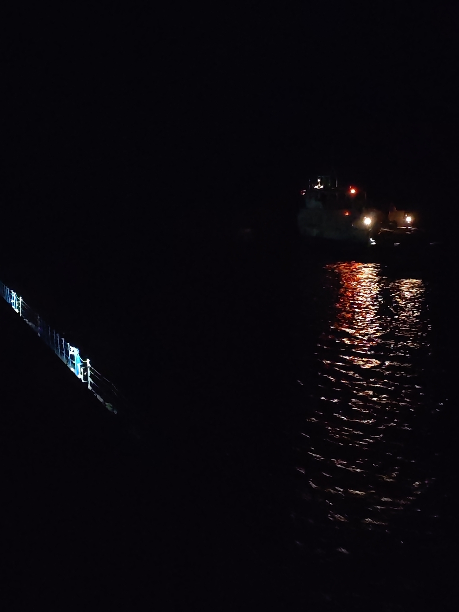 The image depicts a dark nighttime scene, likely on or near water. On the right side, there's a boat or vessel with illuminated lights, which are reflecting on the water's surface. The reflection creates a shimmering, fragmented effect on the water. On the left side of the image, there's a portion of a railing or structure, possibly part of a bridge or another boat. The lighting on the railing is different from the boat lights, suggesting separate light sources. The overall environment is dark, with most of the background obscured by shadows. The location is unknown based on the given information.