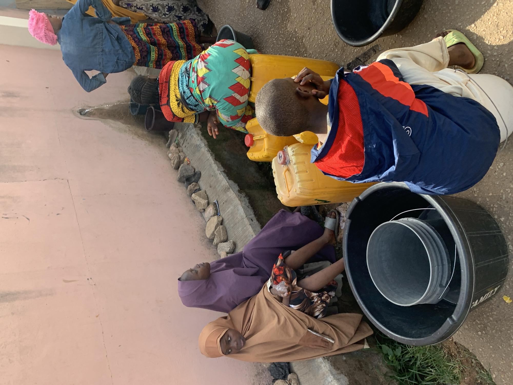 In Jos, Nigeria, several individuals are gathered around a water source, possibly a communal tap or well, to collect water. The setting appears to be a residential or communal area, evidenced by the presence of a pink-painted wall. Individuals are using various containers such as yellow jerrycans, plastic buckets, and possibly smaller pails to store the collected water. One person is wearing a blue and red jacket with white pants, while another is wearing a brightly colored dress. Two women are wearing hijabs, one purple and one brown. The ground area is paved and appears to have a concrete water runoff area. The water source is represented by a small pipe.