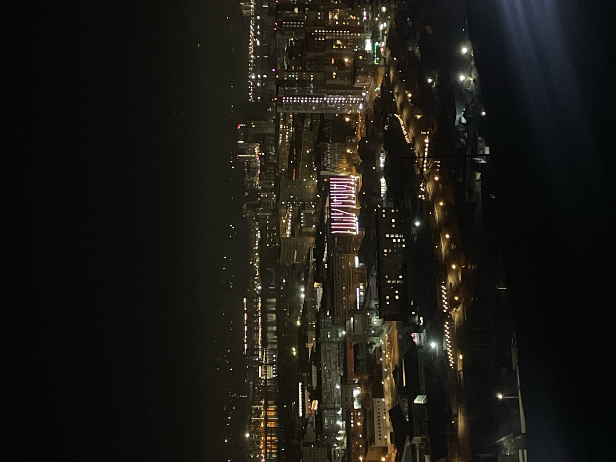 The image shows a nighttime view of Krasnoyarsk, Russia. The cityscape is illuminated with numerous lights from buildings, streets, and possibly vehicles. The left side of the image shows a dark, likely unpopulated area, possibly the Yenisey River. The right side is dominated by city structures, revealing residential and commercial buildings. Some buildings display unique lighting patterns, drawing attention to their architectural features. Streets and pathways are visible as lines of light running throughout the city grid. The image lacks any clear indication of active human presence, but the overall brightness signifies urban activity.