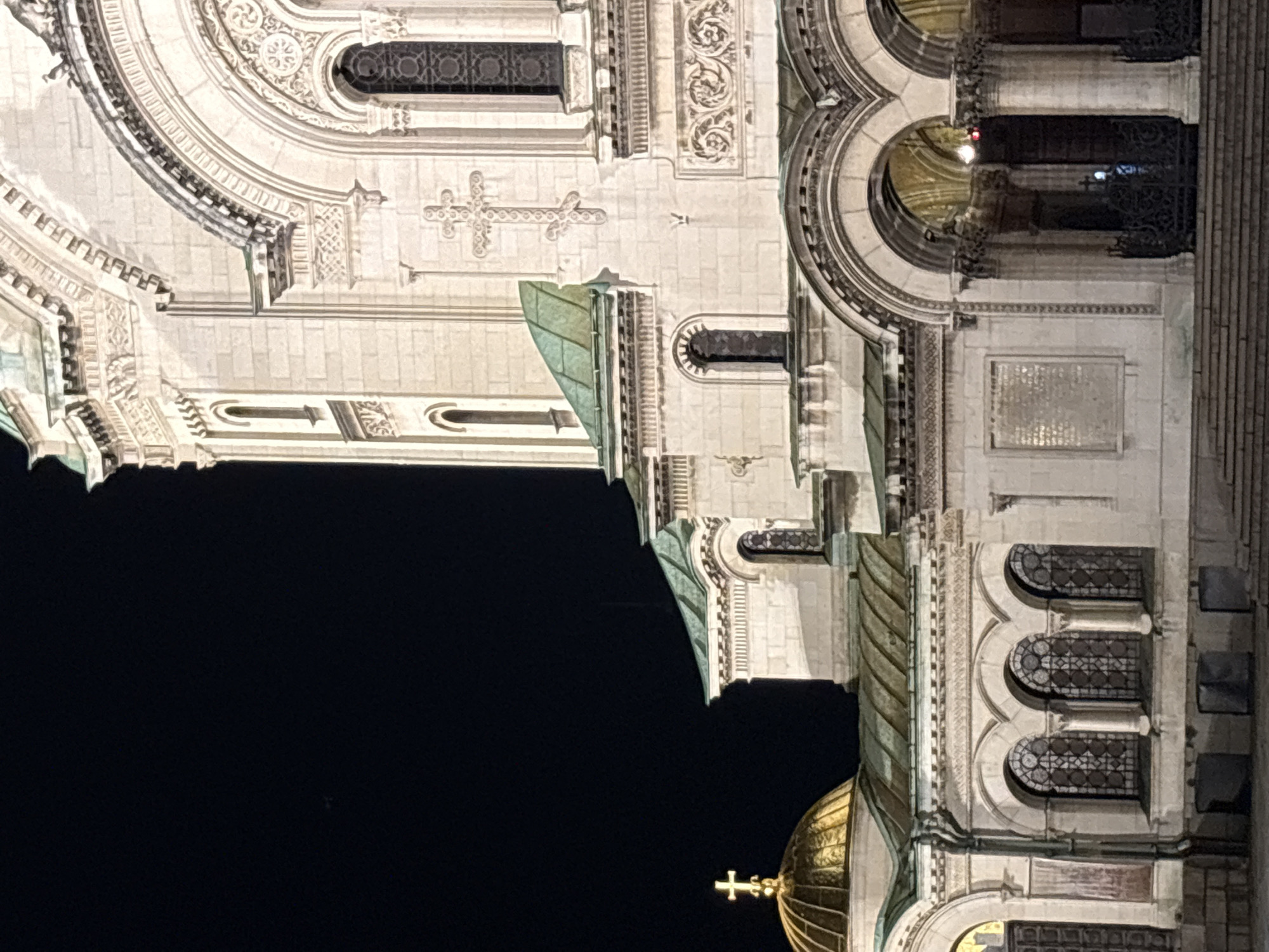 The image shows the Alexander Nevsky Cathedral in Sofia, Bulgaria, at night. The cathedral's architectural details are illuminated, including its golden dome topped with a cross, arched windows, decorative moldings, and crosses carved into the facade. The background is dark, emphasizing the illuminated building.