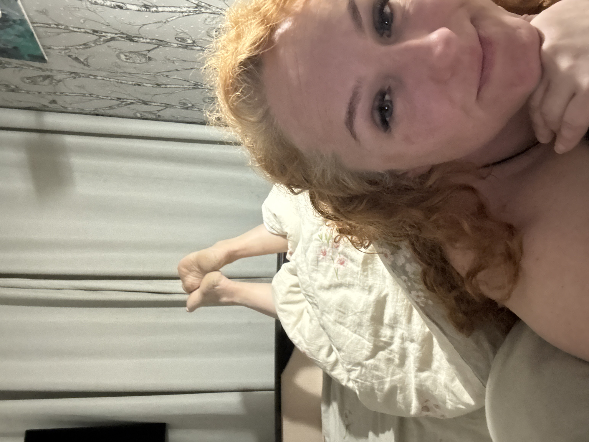A selfie photograph taken indoors shows a fair-skinned woman with red hair and light eyes. She is lying in bed, with a cream-colored floral-patterned bedspread partially visible. Her left foot rests against a gray wall, likely belonging to a bedroom. A television screen is faintly visible at the bottom of the image. The location appears to be in a residential setting, potentially in Chaikovsky, Russia.