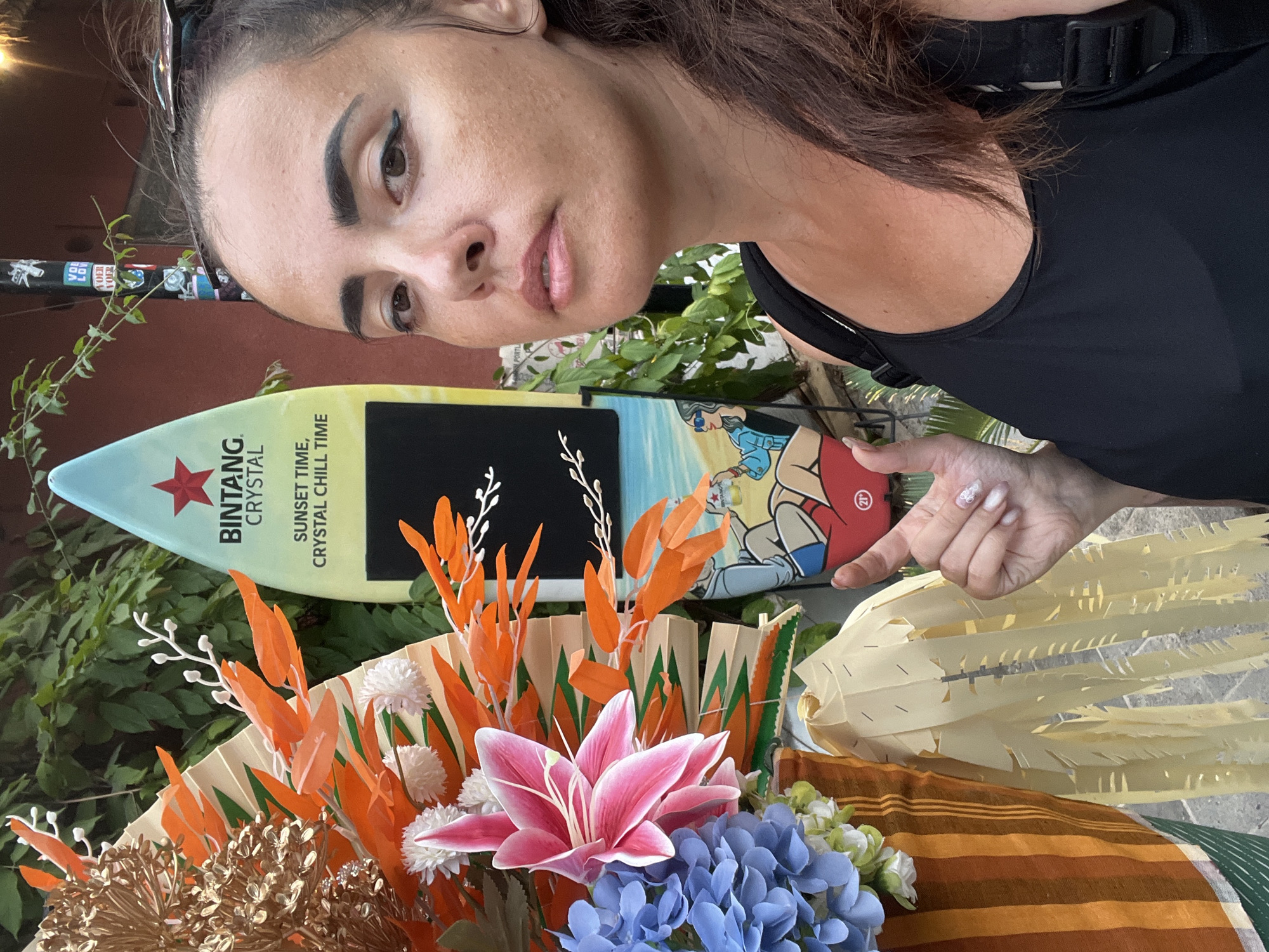 A woman with dark brown hair, wearing a black tank top, is taking a selfie outdoors. She has noticeable eyeliner and her nails are painted with a white pattern. To her left, there is a decorative arrangement featuring a pink lily, blue hydrangeas, and various orange and gold accents, including some fan-like paper elements. Behind the floral display and to the woman's right is a surfboard with the Bintang Crystal logo and the text "SUNSET TIME, CRYSTAL CHILL TIME." The surfboard also features a cartoon illustration of a person sitting by the ocean. The background consists of green foliage and a reddish-brown wall with some stickers on a pole. The scene appears to be in Kuta, Indonesia, possibly near a beachside establishment.