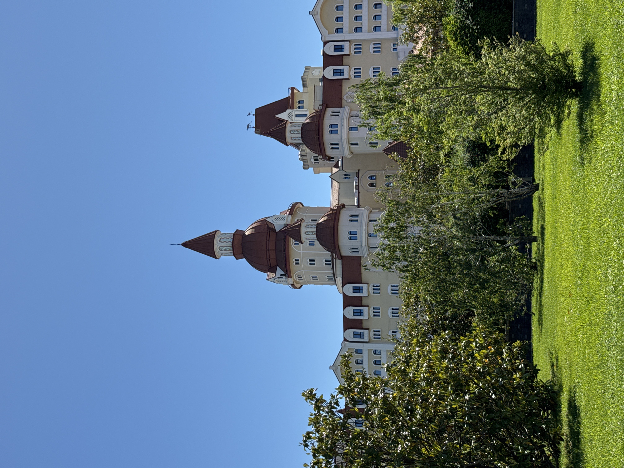 An ornate, multi-story building with distinctive architecture is prominently featured against a clear blue sky. The building, likely a hotel or resort complex, boasts light-colored facades, possibly cream or beige, adorned with numerous windows that are a mix of arched and rectangular shapes, many featuring dark frames. Its most notable features are the multiple towers and turrets, capped with reddish-brown, conical, and rounded roofs. The tallest spires are topped with what appear to be decorative finials or small weathervanes.
In the foreground and to the side of the building lies a lush, vibrant green lawn, meticulously maintained. Several trees with dense green foliage are scattered across the grassy area, casting distinct, elongated shadows, indicating bright sunlight. The sky above is a uniform, cloudless blue.
The time of day is clearly daytime, likely late morning or early afternoon, given the intensity of the sunlight and the clear shadows. The weather is bright and sunny. The architectural style suggests a grand, possibly resort-like, establishment in Sirius, Russia, as per the provided context. There are no people visible, and no specific activity is taking place. No visible text is present in the image.