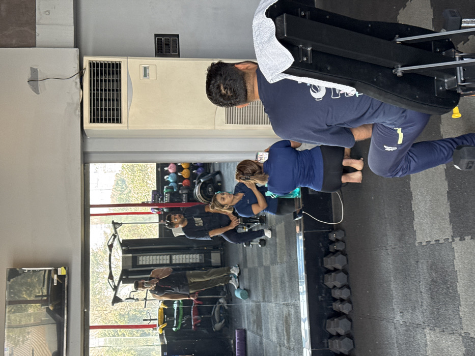 A gym interior is reflected in a large mirror. A woman sits on a weight machine, holding a phone and facing a man in a similar shirt and pants. A second man can be seen reflected further down the mirror, holding a phone in his hand, presumably taking a selfie. Weight racks, dumbbells, and colorful kettlebells are visible in the reflection. A large air conditioner unit sits to the left of the mirror on the wall.