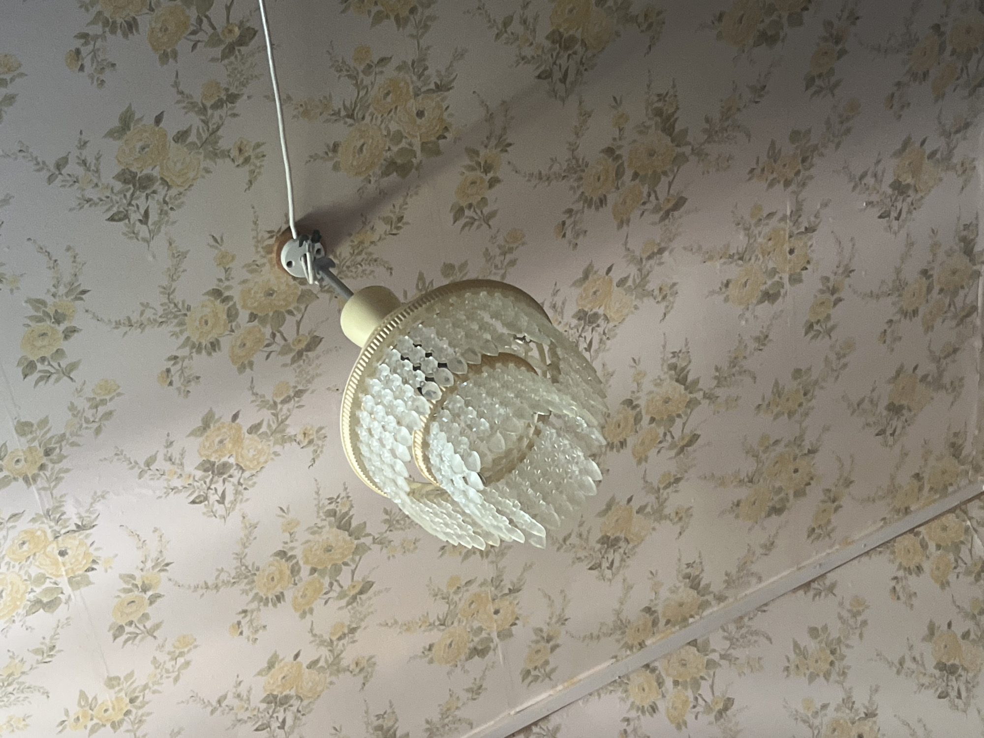 The image depicts a close-up, low-angle view of a chandelier hanging from a ceiling with floral wallpaper. The wallpaper features a pattern of light pink and cream roses with green leaves. The chandelier is a decorative fixture with multiple tiers of clear, faceted glass or acrylic elements that appear to be teardrop-shaped. The chandelier is suspended by a thin rod and is connected to a white electrical fitting on the ceiling. A white electrical cord extends from the fitting. The overall impression is of an interior space, likely a room, with a vintage or traditional aesthetic. There is no indication of the city of Krasnoslobodsk, Russia, in the image.