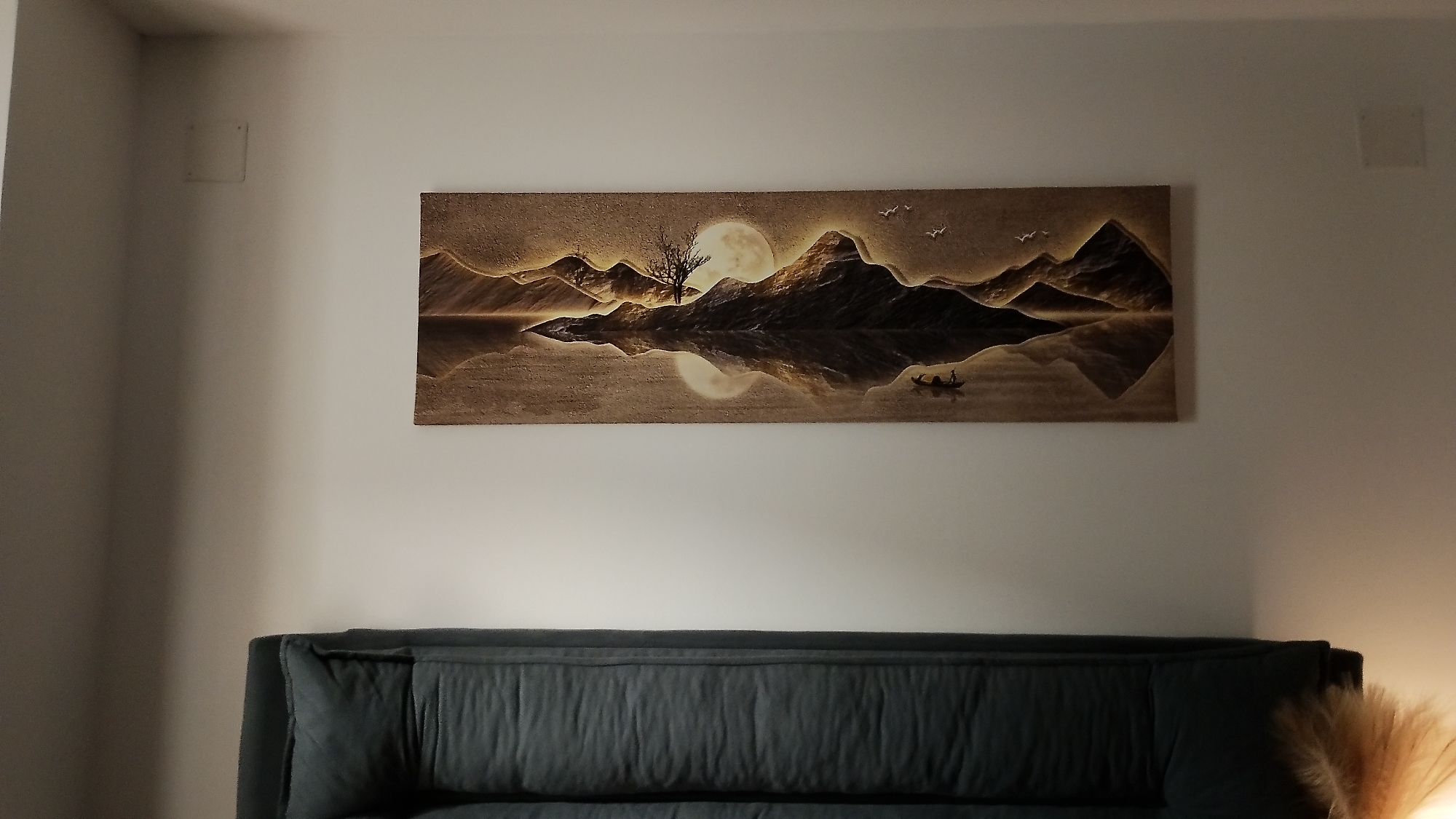 A horizontal, framed artwork depicting a serene mountain landscape at dusk or dawn hangs on a white wall above a dark gray sofa. The artwork features a body of water reflecting mountains, a barren tree, a large moon, and a small boat with two figures. The scene is rendered in warm, sepia tones. Two square white fixtures are visible on the wall, one to the left and one to the right of the artwork. A portion of a floor lamp with a light-colored shade is visible on the far right side of the image, along with a feathery decorative element. The sofa has a plush, tufted backrest. The overall impression is one of a calm, interior living space. The location in the city of Smolyan, Bulgaria is not discernible from the image.