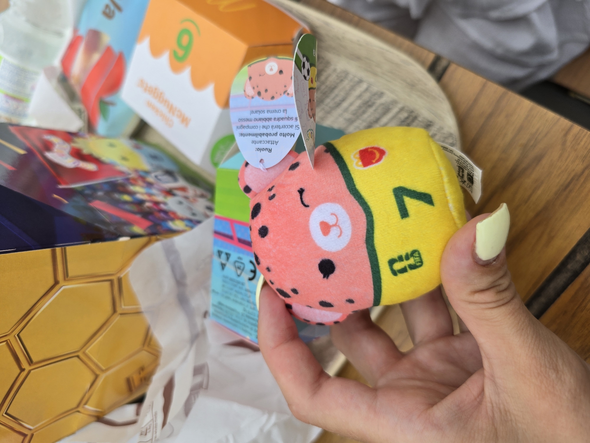 In Fisciano, Italy, a hand with light skin and pale yellow nail polish is holding a pink plush toy with black spots and a yellow band around its middle. The toy has a cartoonish face with closed eyes and a small mouth. A green number "7" is visible on the yellow band, along with a small McDonald's logo. A tag attached to the toy contains Italian text and a drawing of a similar plush character. The background shows a brown wooden surface, a white paper bag with a honeycomb pattern, and partially obscured McDonald's packaging, including a white box with "Chicken McNuggets" and a green number "6" printed on it.