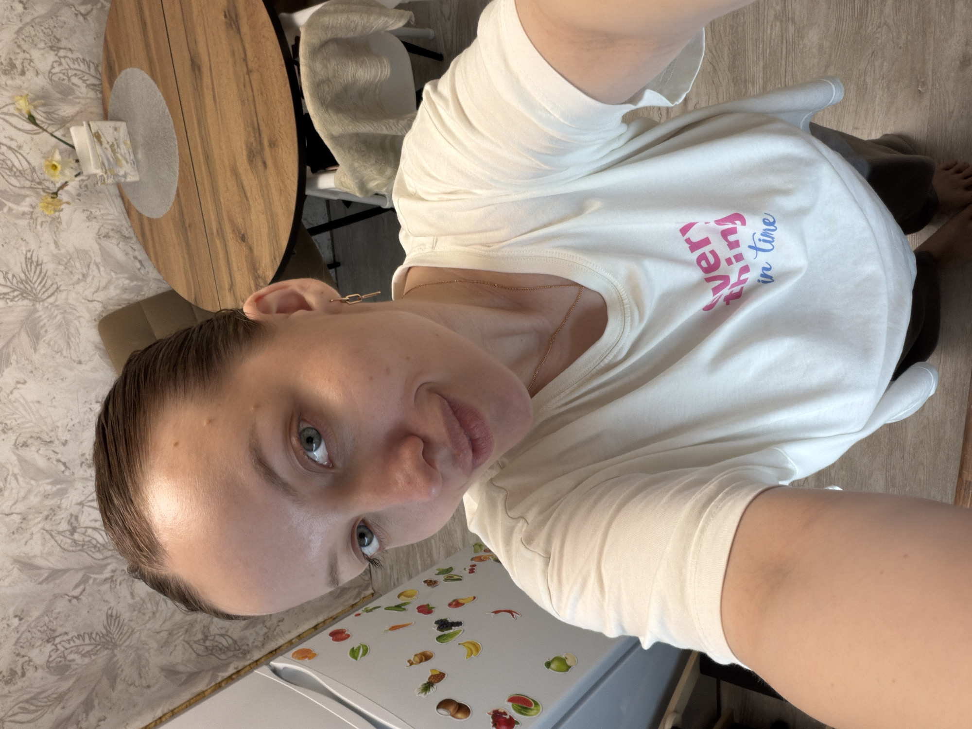 A woman is taking a selfie in what appears to be her home. The background includes a wooden table, a chair, and a refrigerator decorated with fruit magnets. The woman is wearing a white t-shirt with the words "everything in time" printed on it. Part of her legs and feet are also visible. No evidence suggests the location is specifically Reshetnikovo, Russia.