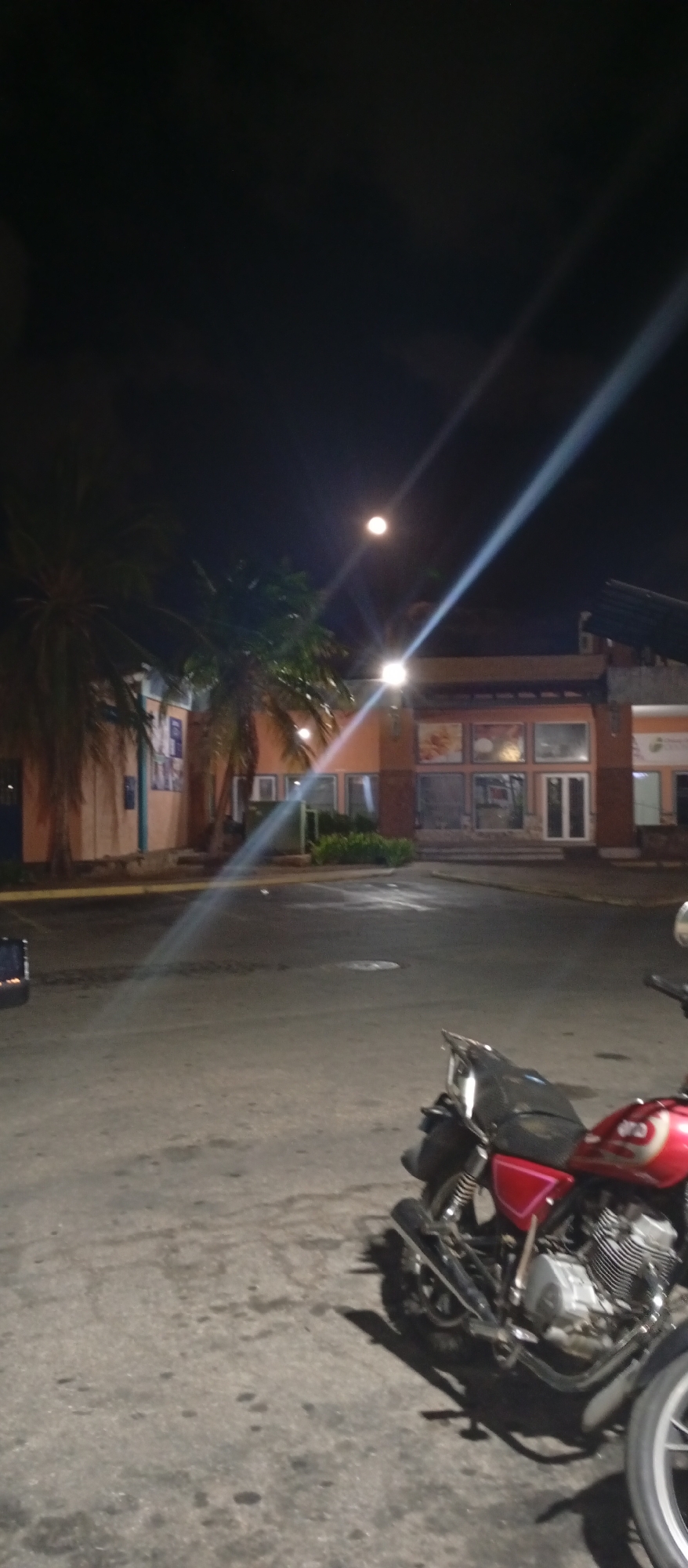 The image shows a dimly lit street scene at night in Juan Griego, Venezuela. A red motorcycle is parked on the asphalt surface in the foreground. Behind the motorcycle, a one-story building is visible with several windows and doors, appearing closed. There are palm trees to the left of the building. A streetlight illuminates the area.