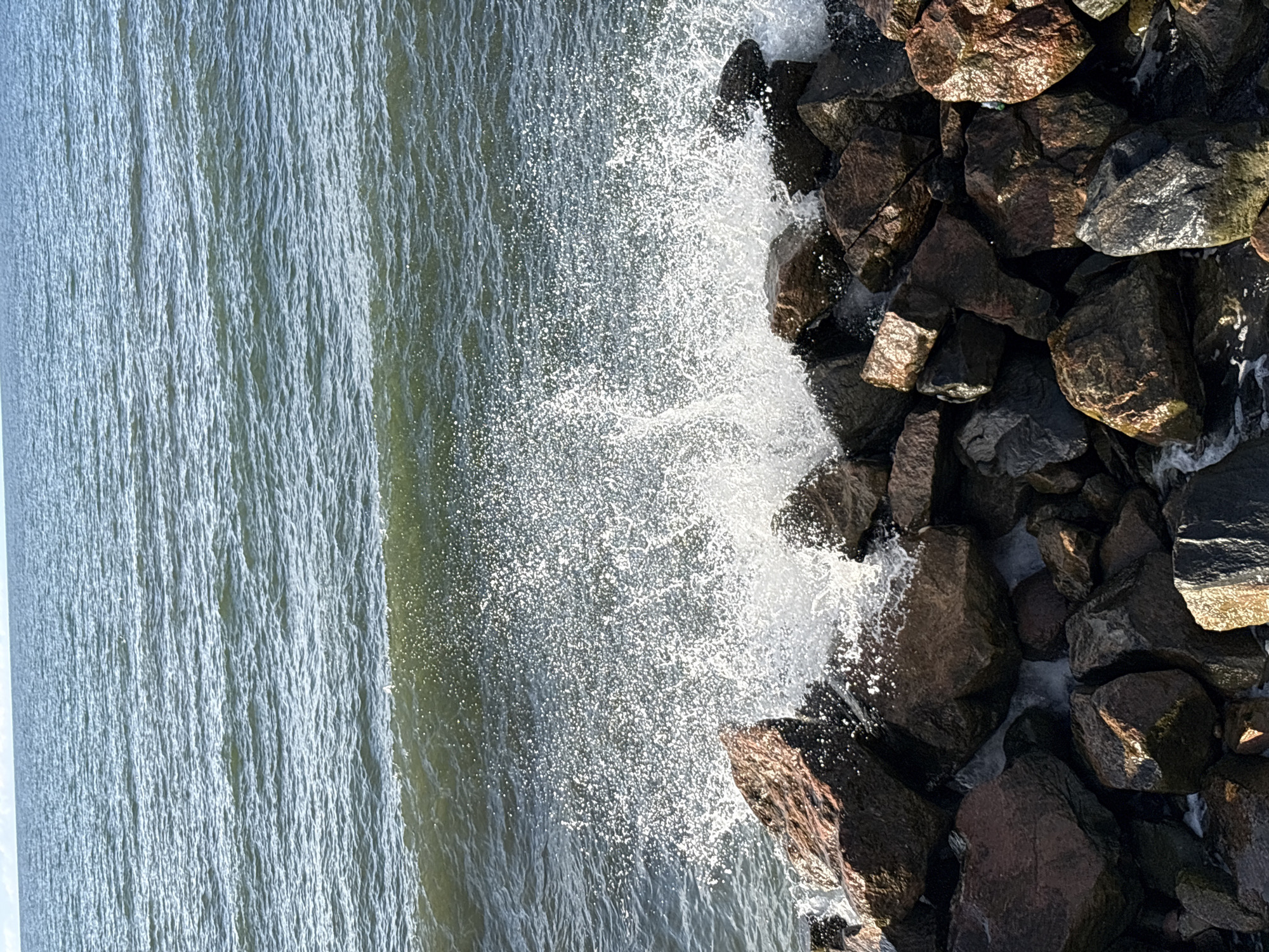 The image captures a close-up view of waves crashing against a rocky shore. The water is a deep, undulating green, with a white spray bursting upwards as it meets the jagged, dark-brown and grey rocks in the foreground. The rocks are piled high, some smooth and rounded, others angular and rough. The texture of the water is captured in its dynamic motion, with small droplets suspended in the air. The horizon line is visible in the distance, separating the water from a pale, hazy sky. This scene is likely located along the coast of Svetlogorsk, Russia, a popular resort town known for its Baltic Sea shoreline and picturesque rocky outcrops. There are no people or animals visible in the image.