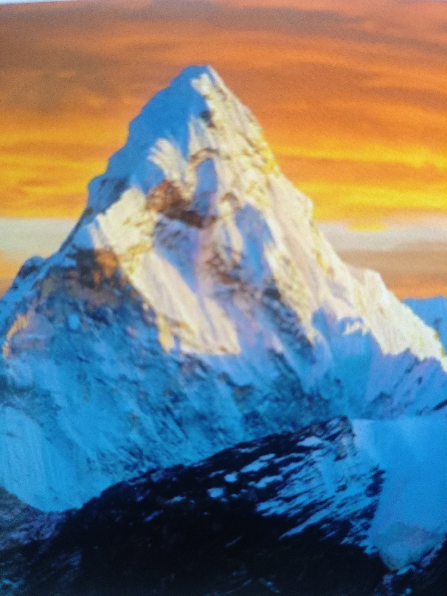 The image shows a painting of a majestic snow-capped mountain against a vibrant orange and yellow sky. The mountain is depicted with sharp, angular features and is partially covered in snow, with visible shadows and highlights that suggest a dramatic lighting effect. The foreground features a darker, rocky terrain also dusted with snow. The style of the painting is somewhat impressionistic, with visible brushstrokes and a rich, textured appearance. There are no people, animals, or man-made structures visible in the image. The painting appears to be a landscape artwork, likely depicting a scene of natural grandeur. The location in Krasnaya Polyana, Russia, is not evident from the visual information in the image itself.