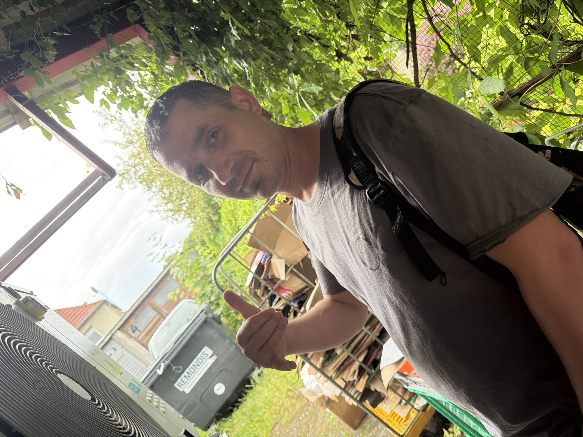 A man in a grey t-shirt gives a thumbs-up gesture while standing in front of a metal cage filled with cardboard boxes. Behind him, a large black dumpster with "REMONDIS" written on it is visible. The background shows a building with a red roof, trees, and foliage. The overall setting appears to be an outdoor area, possibly a recycling or waste disposal site. The man's body language and the presence of sorted materials suggest he is involved in some form of waste management or recycling operation. The scene takes place outdoors, with natural light illuminating the area.