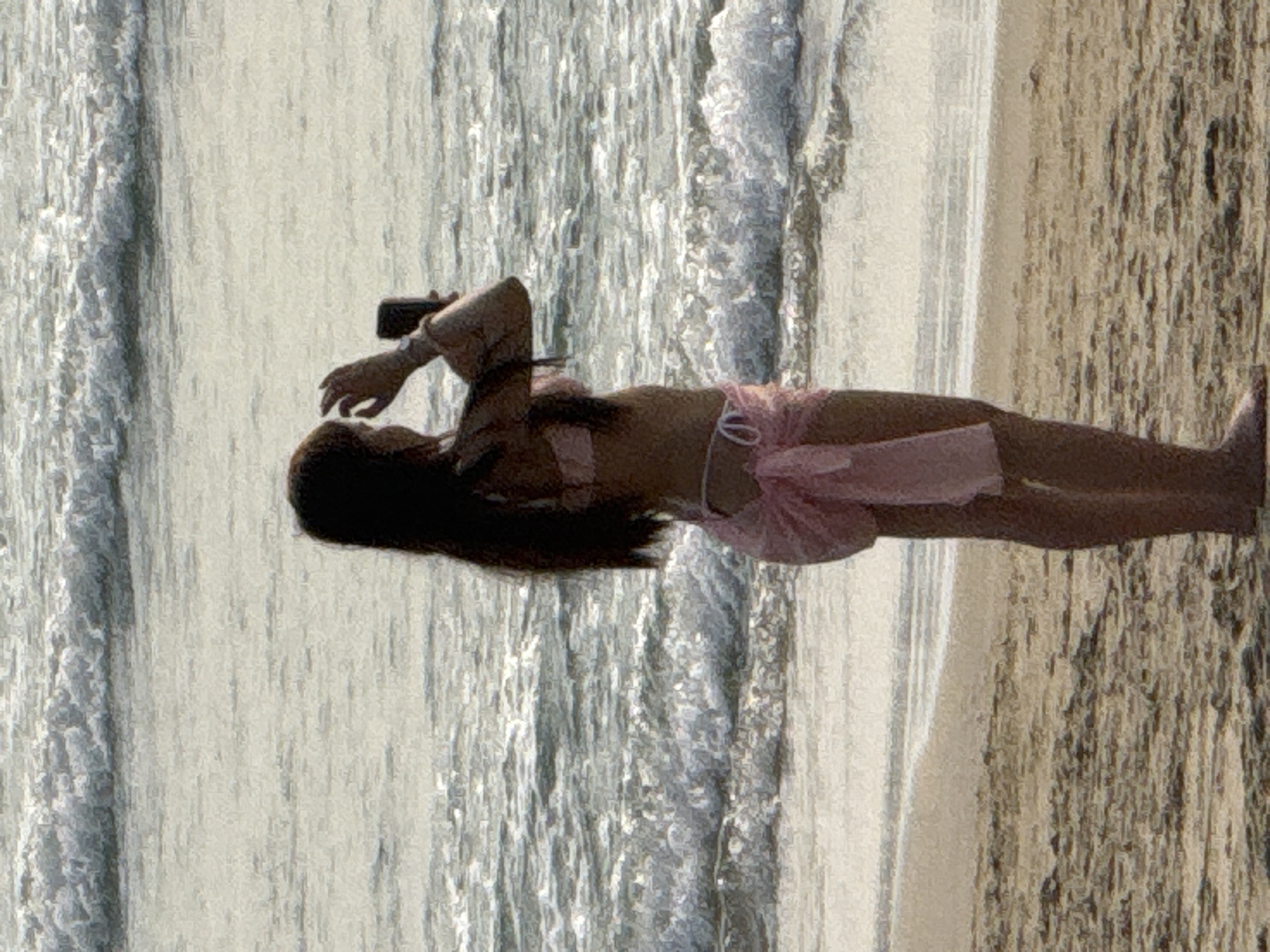 A woman lies on the beach, partially submerged in shallow water. She has long dark hair and is wearing a pink two-piece bathing suit. She is holding a phone or small camera in her hand. The beach is sandy, and waves are breaking nearby. Based on the image and its likely origin, the location is potentially one of the beaches in the city of Đà Nẵng, Vietnam.