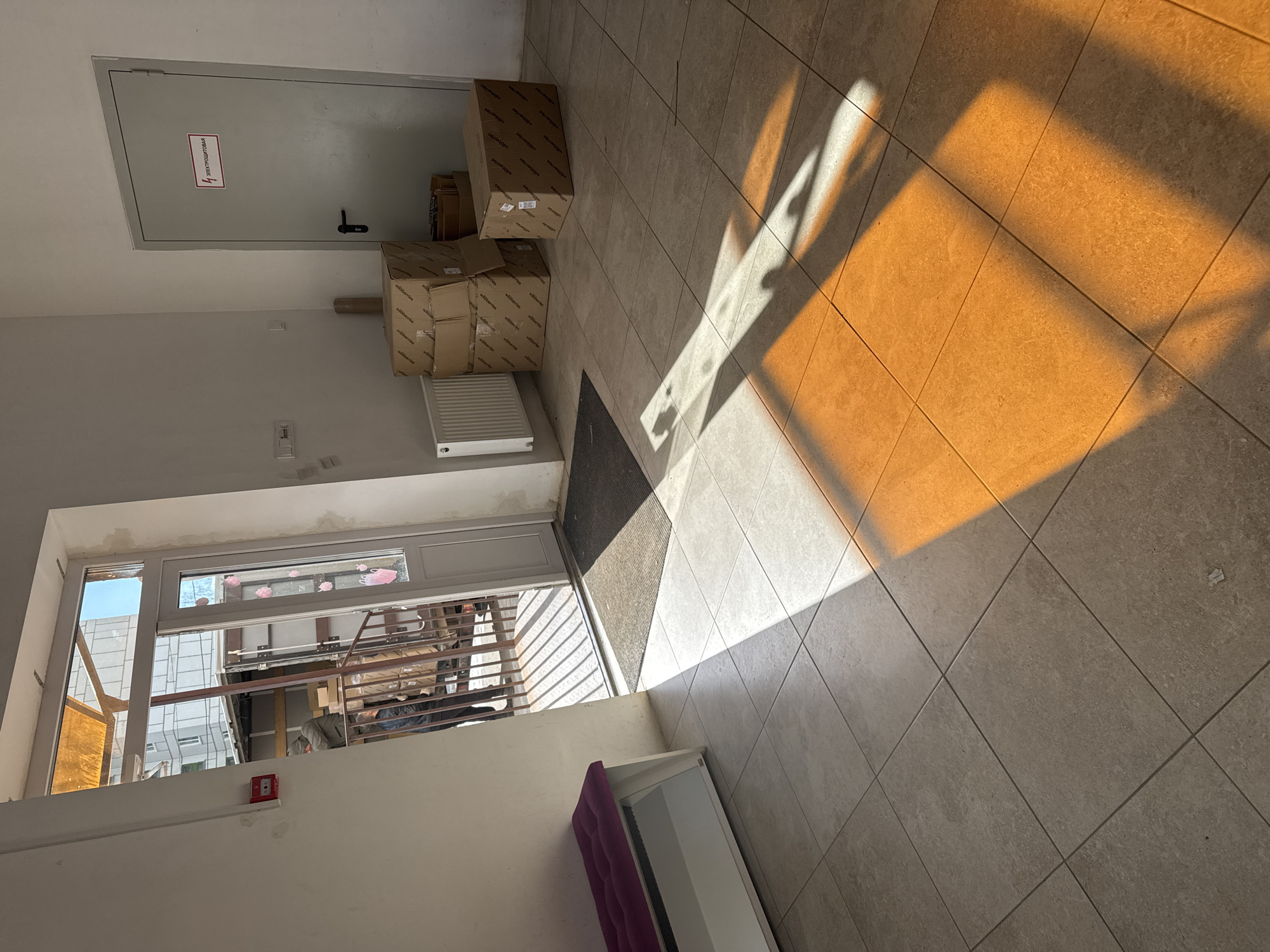 The image depicts an indoor hallway or common area within a building, characterized by light-colored walls and a light-toned tiled floor. Strong sunlight enters from the right, casting long, well-defined shadows across the floor, indicating either early morning or late afternoon.
On the left side of the frame, a grey metal door is visible, featuring a black handle. A rectangular red and white sign on this door clearly states "ЭЛЕКТРОЩИТОВАЯ," which translates from Russian as "ELECTRICAL PANEL ROOM" or "ELECTRICAL SWITCHBOARD ROOM." Below and to the right of this door, several cardboard boxes are stacked. These boxes prominently display the logo "wildberries," identifying them as packages from the popular Russian e-commerce retailer. A white, wall-mounted radiator is situated beneath these boxes.
Further along the hallway on the left, a large window or glass door allows a view of an exterior or an adjacent area, revealing parts of a modern building facade and metal railings. A red fire alarm device is affixed to the wall just below this window. In the bottom right foreground, a small white cabinet or bench with a purple upholstered top is partially visible.
No people are present in the scene, and no specific activity is taking place other than the implied storage or recent delivery of the Wildberries packages. The setting appears functional and utilitarian. Given the Russian text and the Wildberries branding, the location context of Chaikovsky, Russia, is consistent with the visual evidence.
