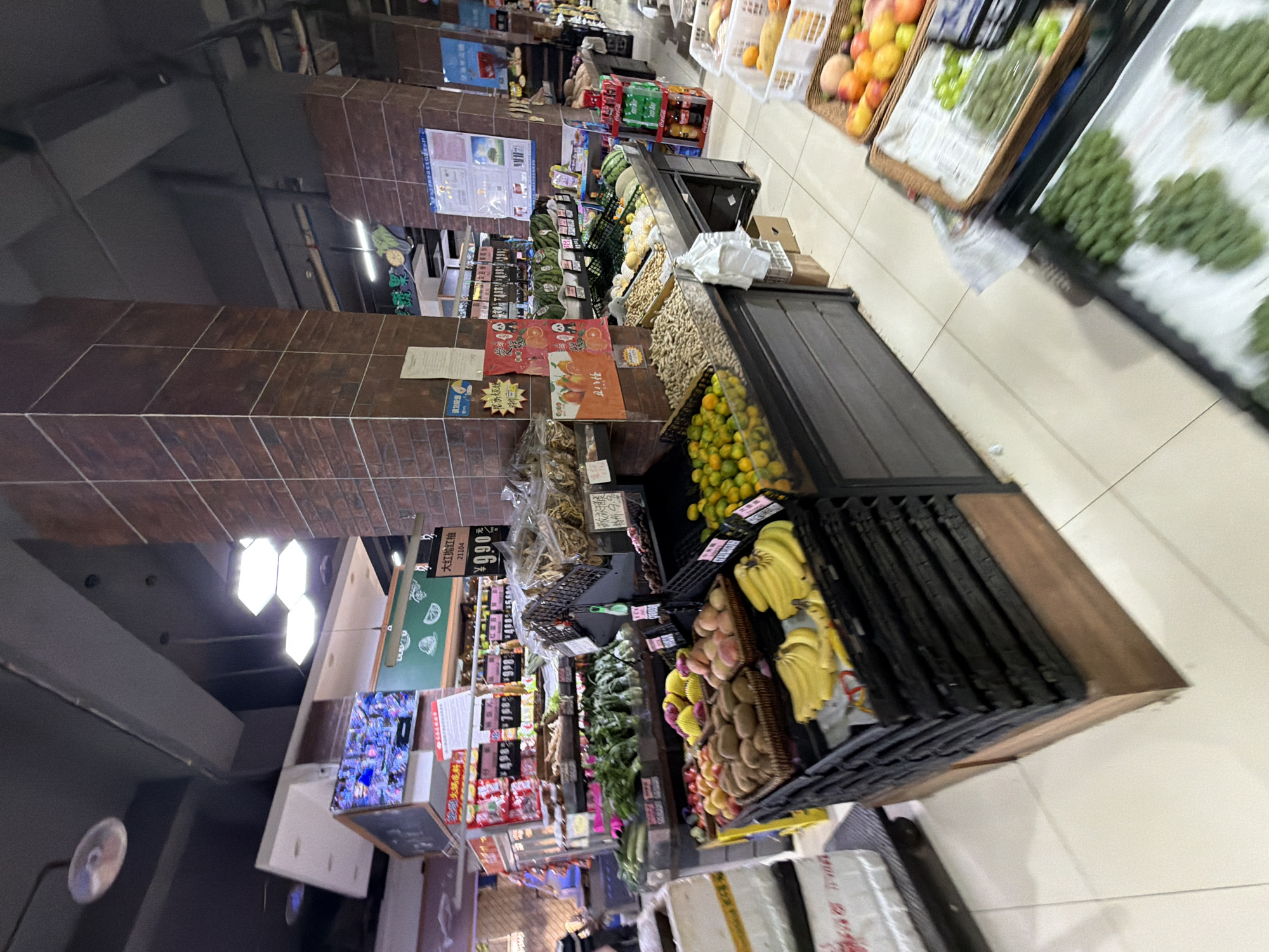 The image depicts a grocery store produce section, likely located in the city of Jiangjin, China, given the presence of Chinese text on signage. The scene is well-lit with overhead fluorescent lighting and showcases a variety of fresh fruits and vegetables displayed in black plastic crates and wicker baskets. Prominently featured are bananas, kiwis, potatoes, apples, oranges, and what appear to be small tangerines or kumquats. Some produce is bagged in clear plastic, while others are loose. Behind the fresh produce, shelves are stocked with packaged goods and more vegetables. A large brick pillar is a central structural element in the foreground. Several signs with Chinese characters are visible, indicating product information and pricing. The floor is tiled in a light beige color. There is a partial view of a person on the left side of the image, seemingly a shopper, interacting with the merchandise. The overall environment suggests a typical supermarket setting focused on fresh food retail.