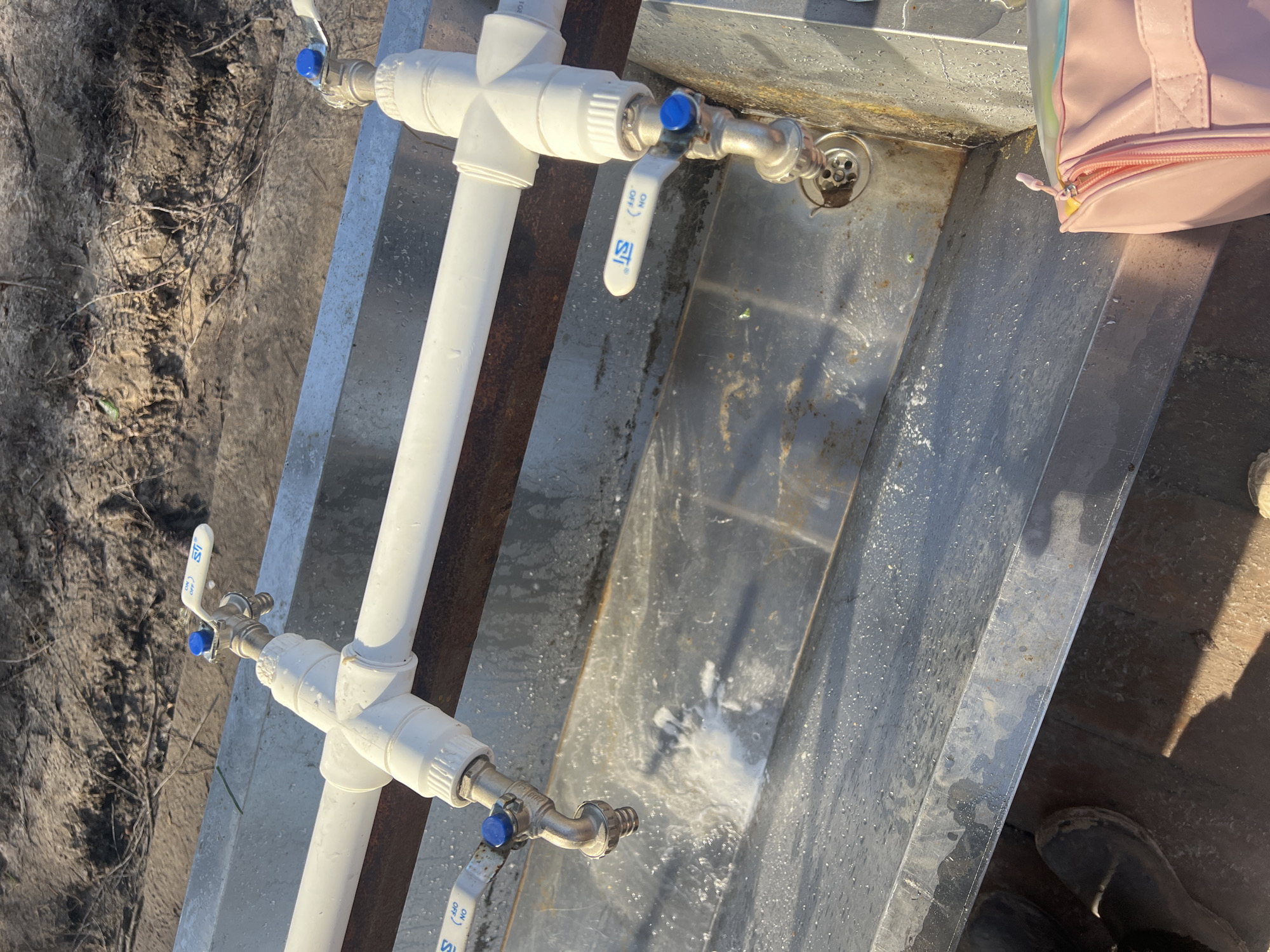 The image depicts an outdoor, utilitarian wash station, likely situated in a rural or garden environment consistent with Lesnoye, Russia. The scene is brightly lit by direct sunlight, casting sharp shadows and highlighting water droplets, indicating a clear, sunny day.
At the center, a plumbing system is visible, constructed from white plastic pipes arranged in a vertical and horizontal configuration. Two silver-colored faucets, each fitted with a blue-handled ball valve, are connected to this piping. The lower faucet is actively running, with water splashing onto a rectangular, worn metal trough or sink below it. This trough exhibits signs of use, including water splashes and minor discoloration, and features a drain with a grate in its upper left section.
To the right of the metal trough, a light pink bag with a visible zipper is present, suggesting human activity. In the bottom right corner, part of a dark boot or shoe is visible on a darker, possibly wooden, surface. The background consists of dry, uneven ground, characterized by sandy soil and sparse, withered vegetation and twigs.
The handles of the ball valves clearly display the text "ON" and "OFF" in blue, along with what appears to be the manufacturer's logo, "STI," also in blue. While no individuals are directly pictured, the objects within the frame imply recent or ongoing human presence and activity related to water usage or washing. The overall impression is one of a functional, basic setup in a natural, rustic setting.