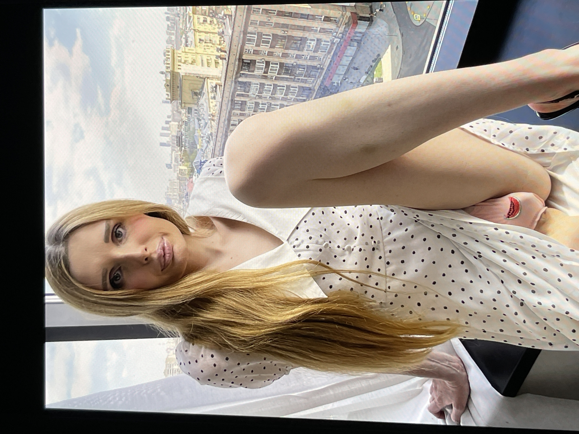 The image displays a woman with long blonde hair and light-colored eyes, wearing a white polka dot dress. She is seated indoors, with a city skyline visible through a large window in the background. The lighting suggests it is daytime, possibly late afternoon, with natural light illuminating the scene. The woman is posing with one leg raised, and her facial expression appears neutral or slightly amused. A pair of black high-heeled shoes are visible on her feet, and she is wearing pink socks with a watermelon design. Her hands are resting on her lap. The overall setting appears to be a modern apartment or hotel room with a view.