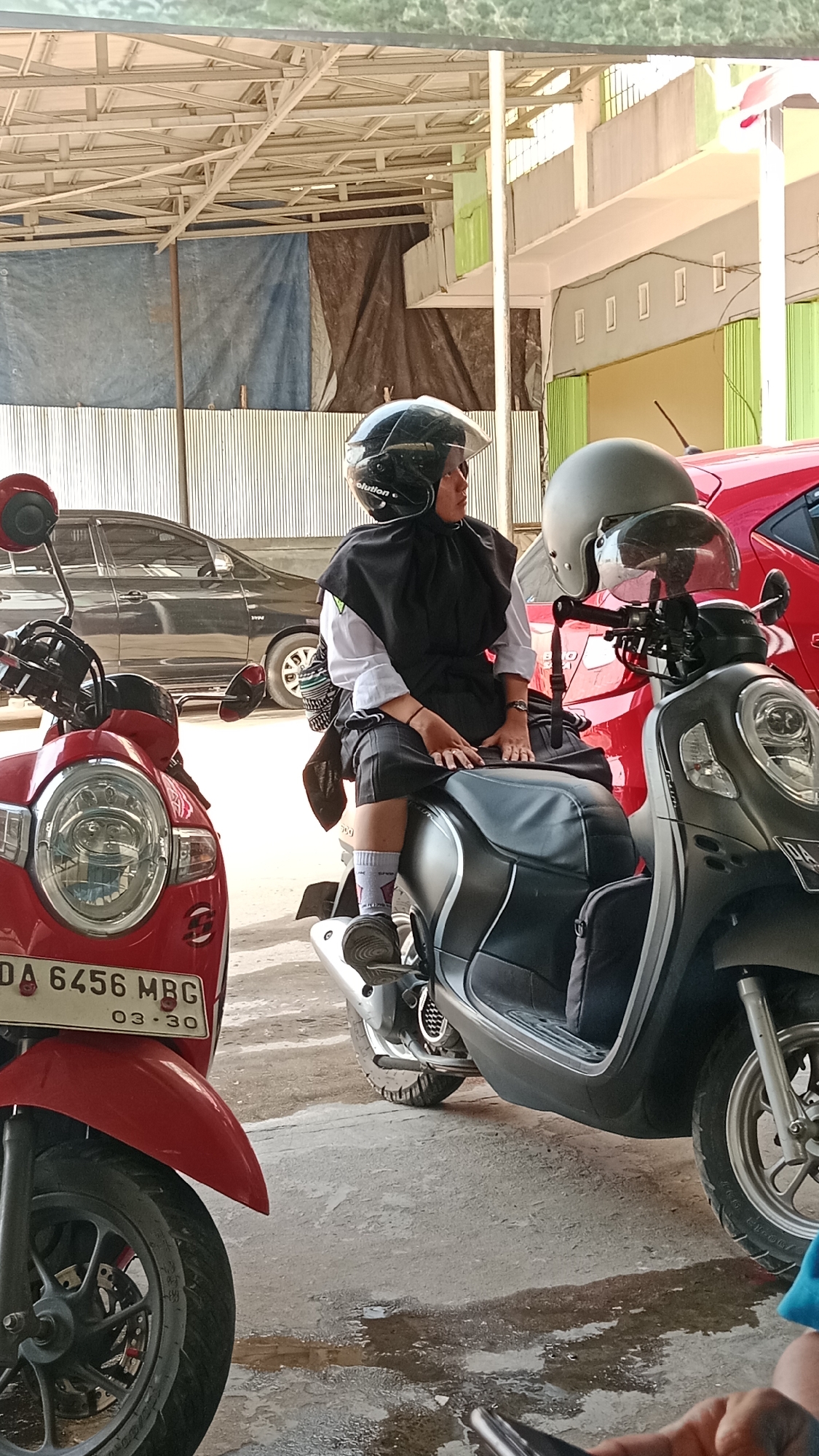 A young woman wearing a black headscarf, white shirt, and dark shorts, sits on a dark gray scooter. She has a black helmet on her head, and another gray helmet is visible on the handlebars of the scooter. A red scooter is parked to her left. In the background, there is a dark gray car, a building with green siding and a light-colored roof, and a tarp draped over a structure. The ground is a concrete surface with some wet patches. The scene appears to be an outdoor parking area. The license plate of the red scooter reads "DA 6456 MBG 03.30". The license plate of the gray scooter is partially visible, showing "DA". This scene is located in Handil Bakti, Indonesia.