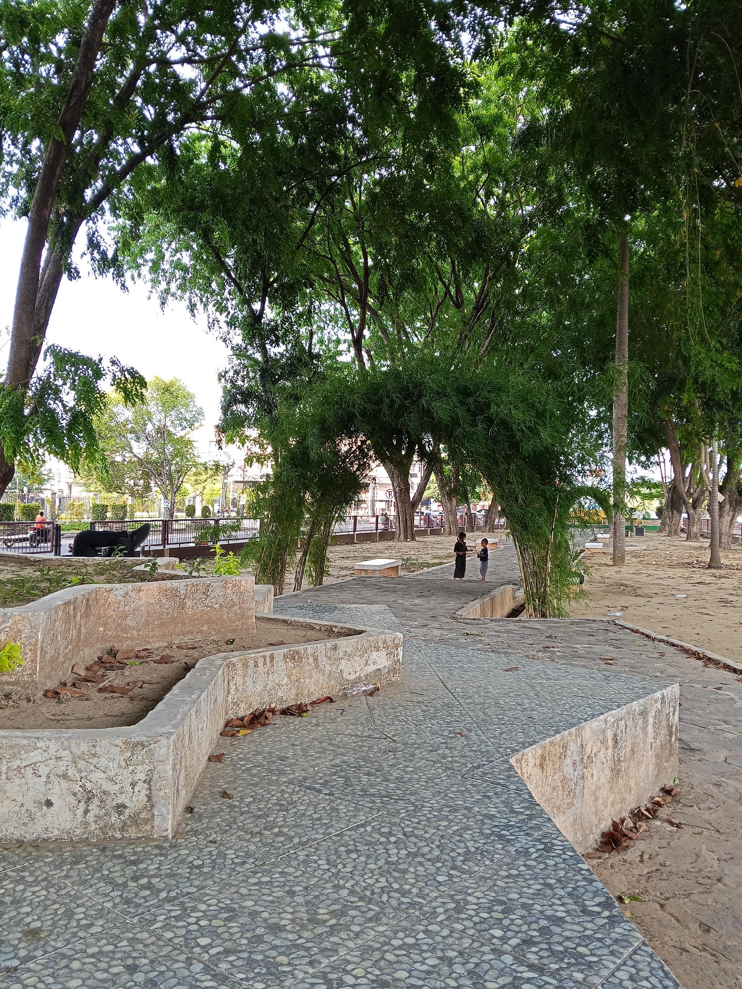 This image depicts a serene outdoor scene in a public park or garden in Lhokseumawe, Indonesia. The setting is dominated by numerous large, mature trees with dense green foliage, providing natural shade across the area. A winding pathway, paved with small, gray and white pebbles, runs through the foreground and middle ground.
On the left side of the pathway, there are low, rustic concrete planters or low walls, some containing dry leaves. Behind these, a dark, animal-shaped sculpture (resembling a tapir) is visible on a raised platform. Further back, a black metal fence separates the park from what appears to be a street or urban area, where buildings and additional trees can be seen. A person on a bicycle is partially visible beyond the fence, and a distant circular road sign with a red border and a white symbol is faintly discernible.
In the center-right of the image, two small children are standing on the paved path. One child wears a dark, possibly traditional, long garment, while the other is dressed in a light-colored top and shorts. They are positioned near a unique archway or tunnel-like structure formed by thick, green bamboo plants growing over the pathway. The ground bordering the path is mostly sandy or dirt.
The scene is captured during the daytime under bright, clear conditions, with sunlight filtering through the tree canopy. The overall atmosphere is peaceful and calm, suggesting a typical day in a community park.