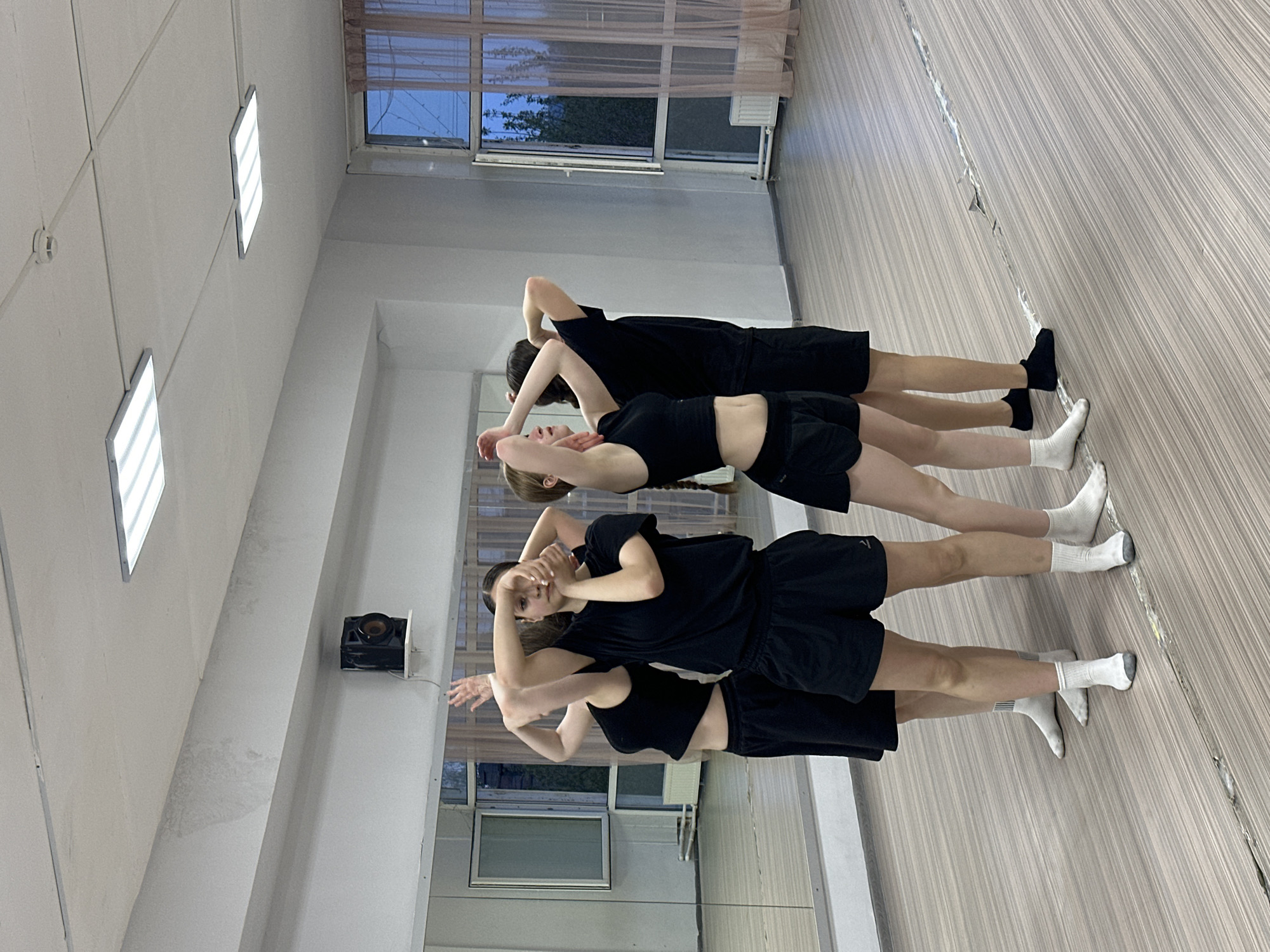 The image shows three people in a dance studio in Votkinsk, Russia. They are lined up in a row, dressed in black athletic wear (shorts and shirts/tank tops) and white socks. The studio has a wooden floor and a wall covered in mirrors. Natural light is coming through a window. There is a projector mounted on the ceiling.